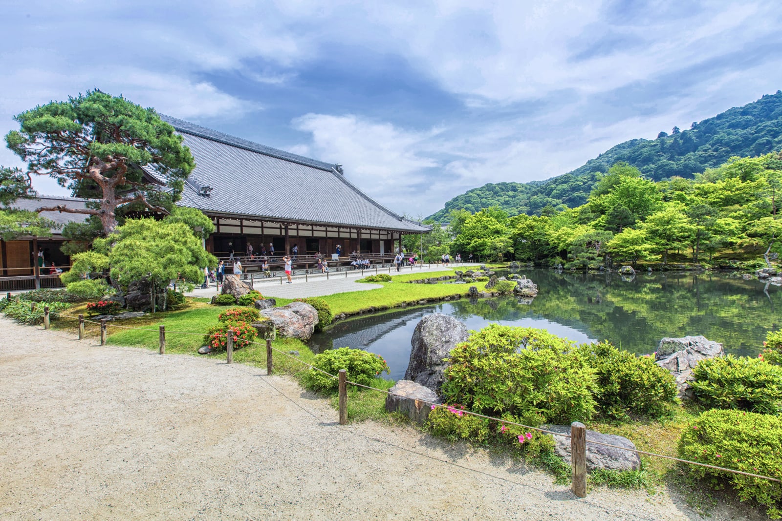 Tenryu-ji's pond garden with rocks and clipped shrubs, a long tiled-roof hall on the left and green hills behind.