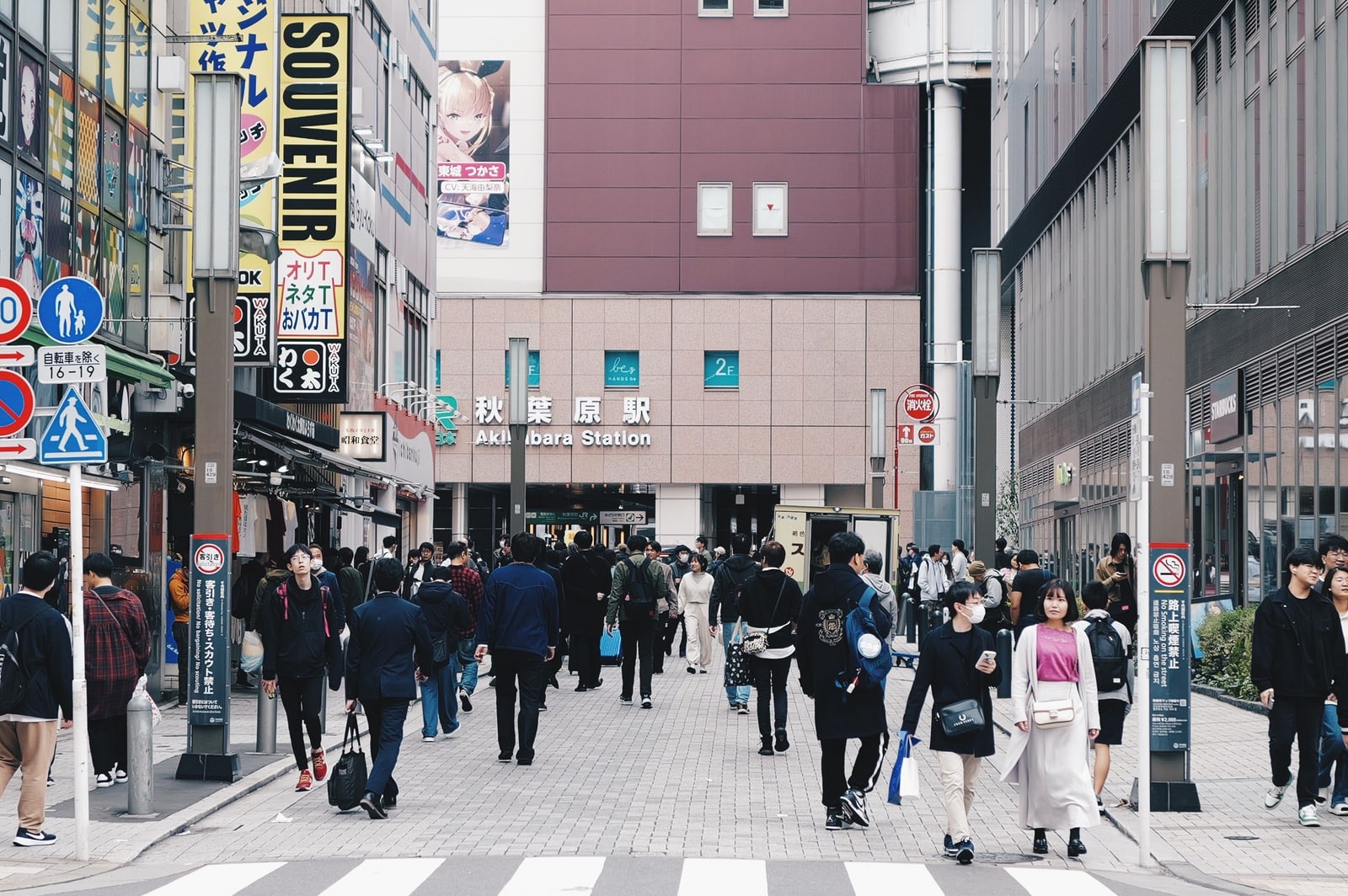 A paved street in daylight leading to the Akihabara Station building, pedestrians walking both ways