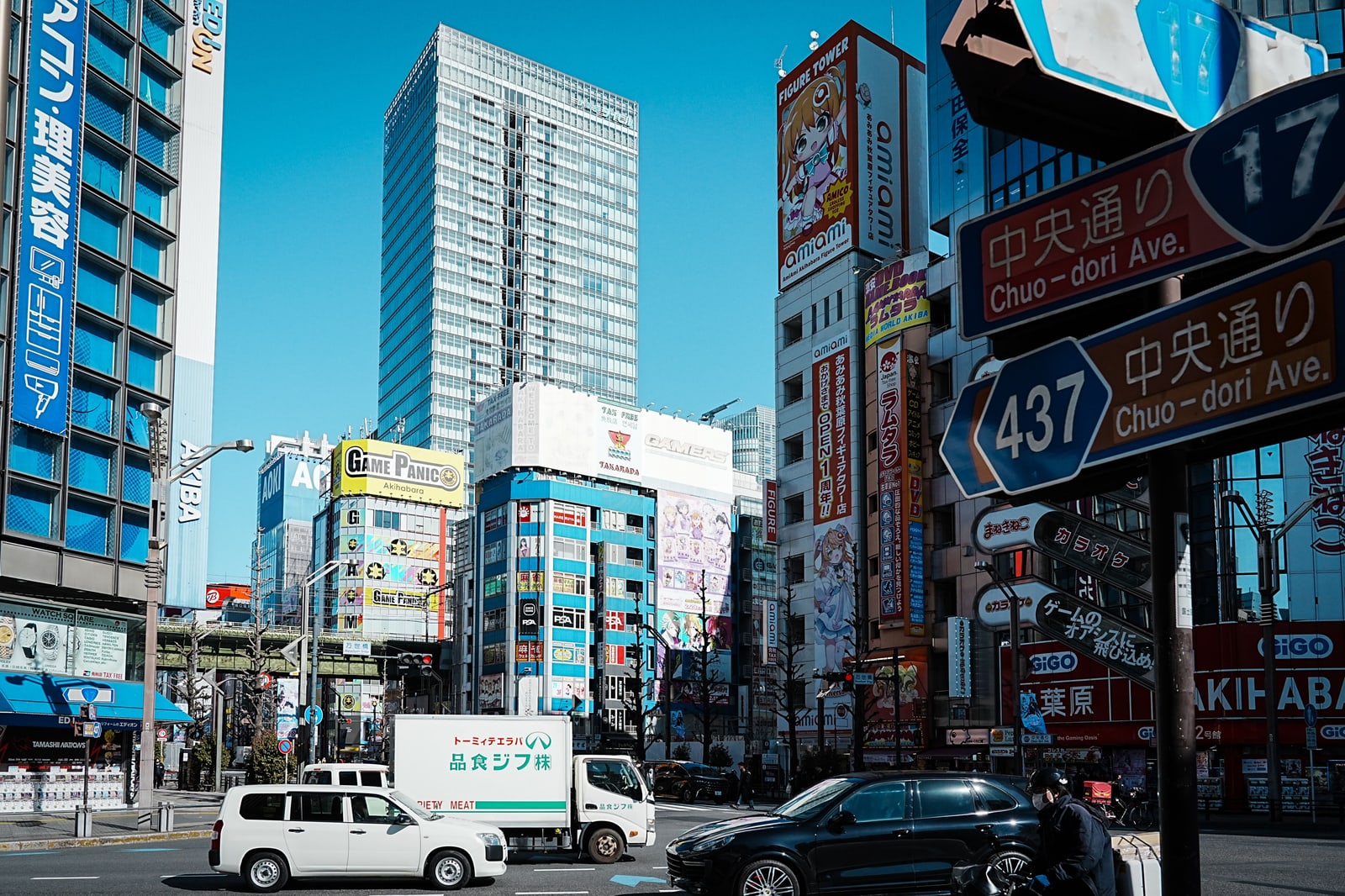 A crossing on Chuo-dori in Akihabara by day under a blue sky, with blue road signs reading 中央通り Chuo-dori Ave., a white van, a truck and a black car, and shop towers behind