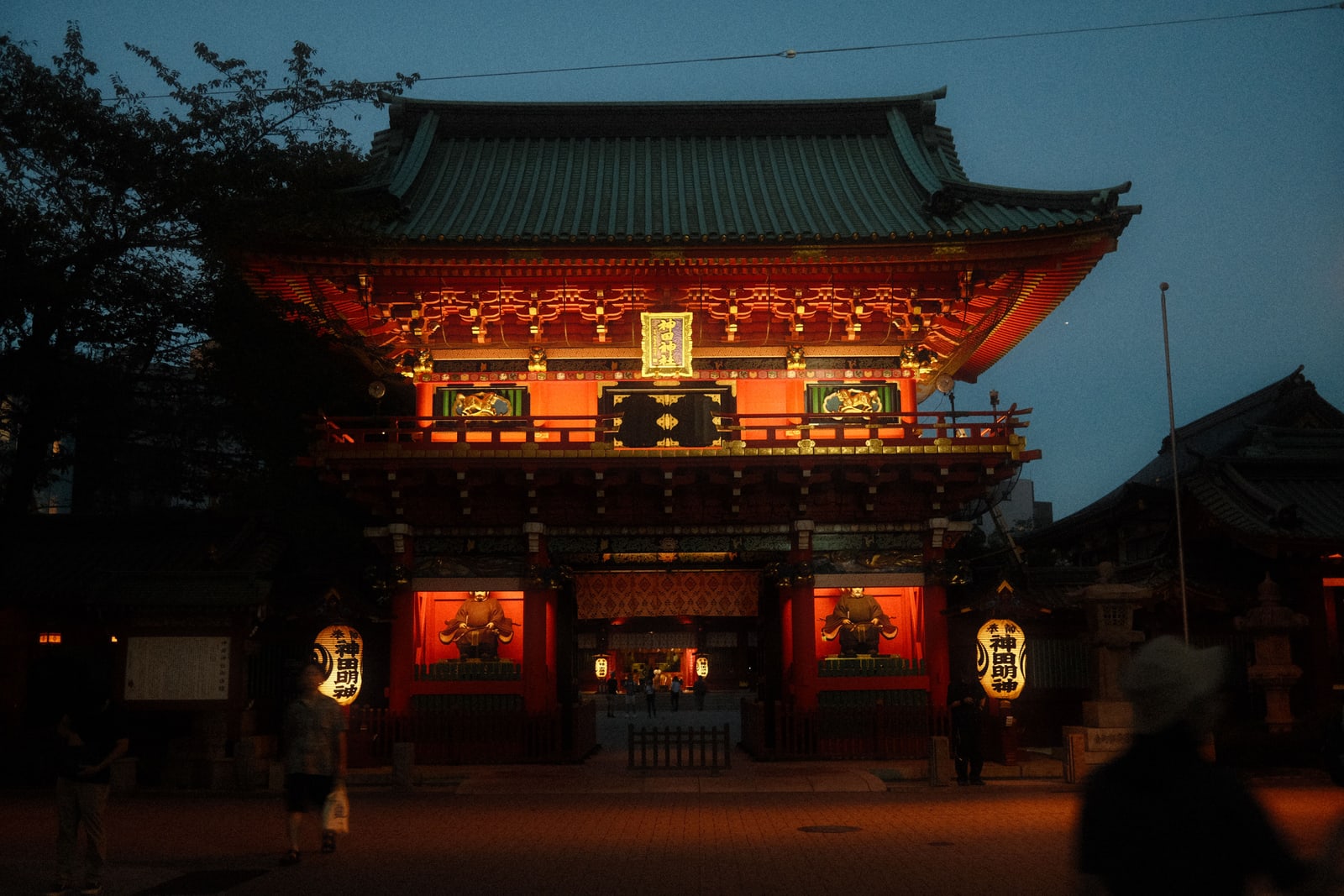 The lit red main gate of Kanda Myojin at dusk with its green tiled roof and two large paper lanterns lettered 神田明神
