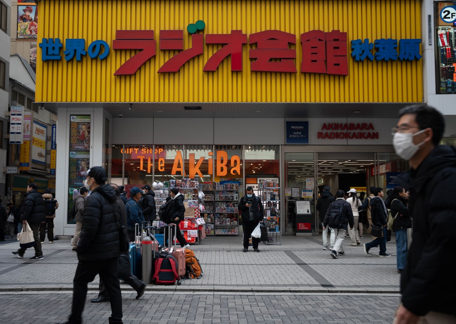 The yellow-fronted Akihabara Radio Kaikan on a brick-paved pedestrian street, with people walking past and a few suitcases at the kerb
