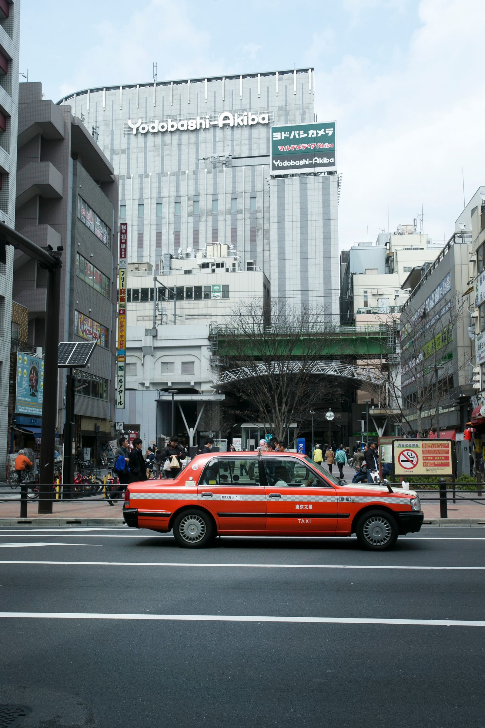 The grey tower of Yodobashi-Akiba with its green Yodobashi Camera sign, seen over a road with a red taxi and a rail viaduct in front