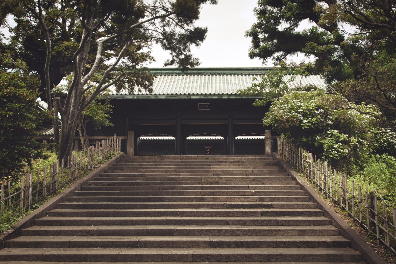 A wide stone staircase rising between trees and bamboo fences to a dark gate with a green tiled roof