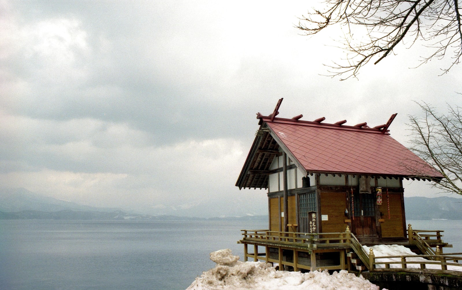 A small red-roofed shrine on a raised deck at a snowy lakeshore, grey water and hills beyond