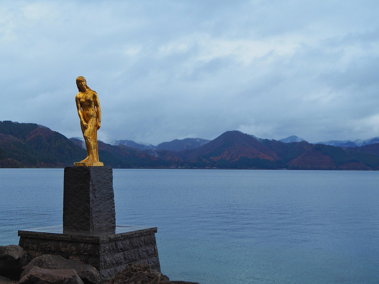 A gold statue on a stone plinth at the edge of a wide lake, autumn hills and low cloud beyond