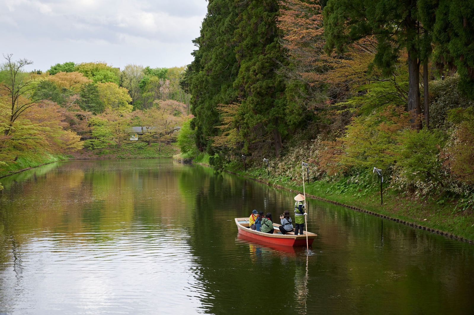 A boatman poling a red wooden boat with passengers along a moat in Hirosaki Park, spring trees on the banks