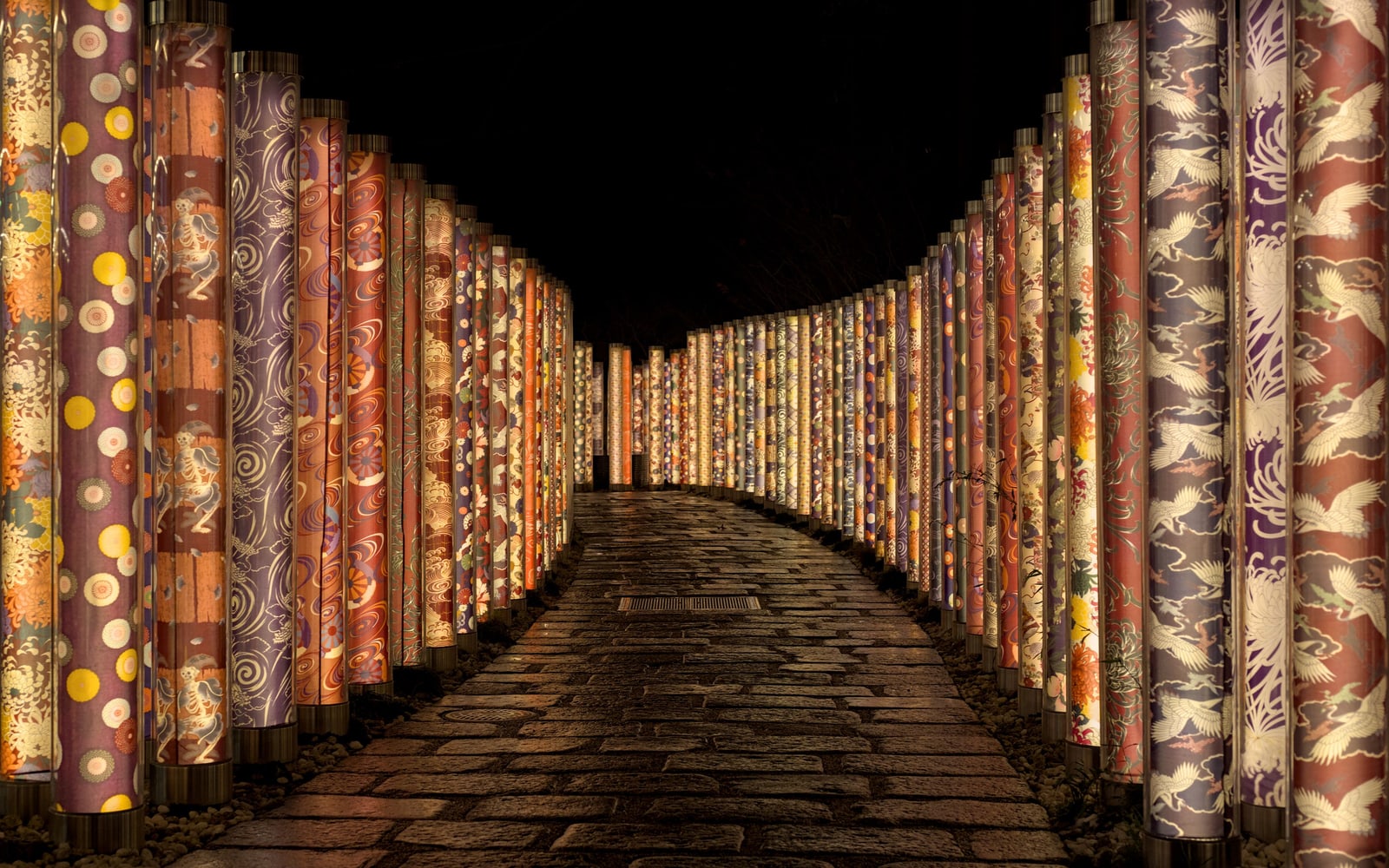 A curving cobbled lane at night between two rows of tall poles wrapped in colourful kimono-pattern fabric and lit from within