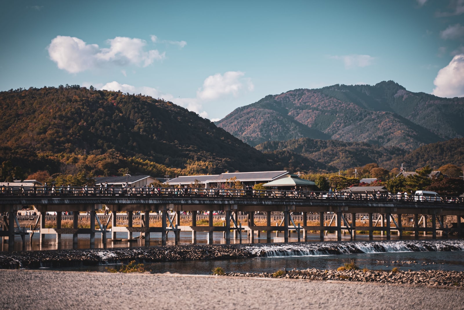 The long timber Togetsukyo bridge across the Katsura river in Arashiyama, autumn-colored hills behind it under a blue sky.