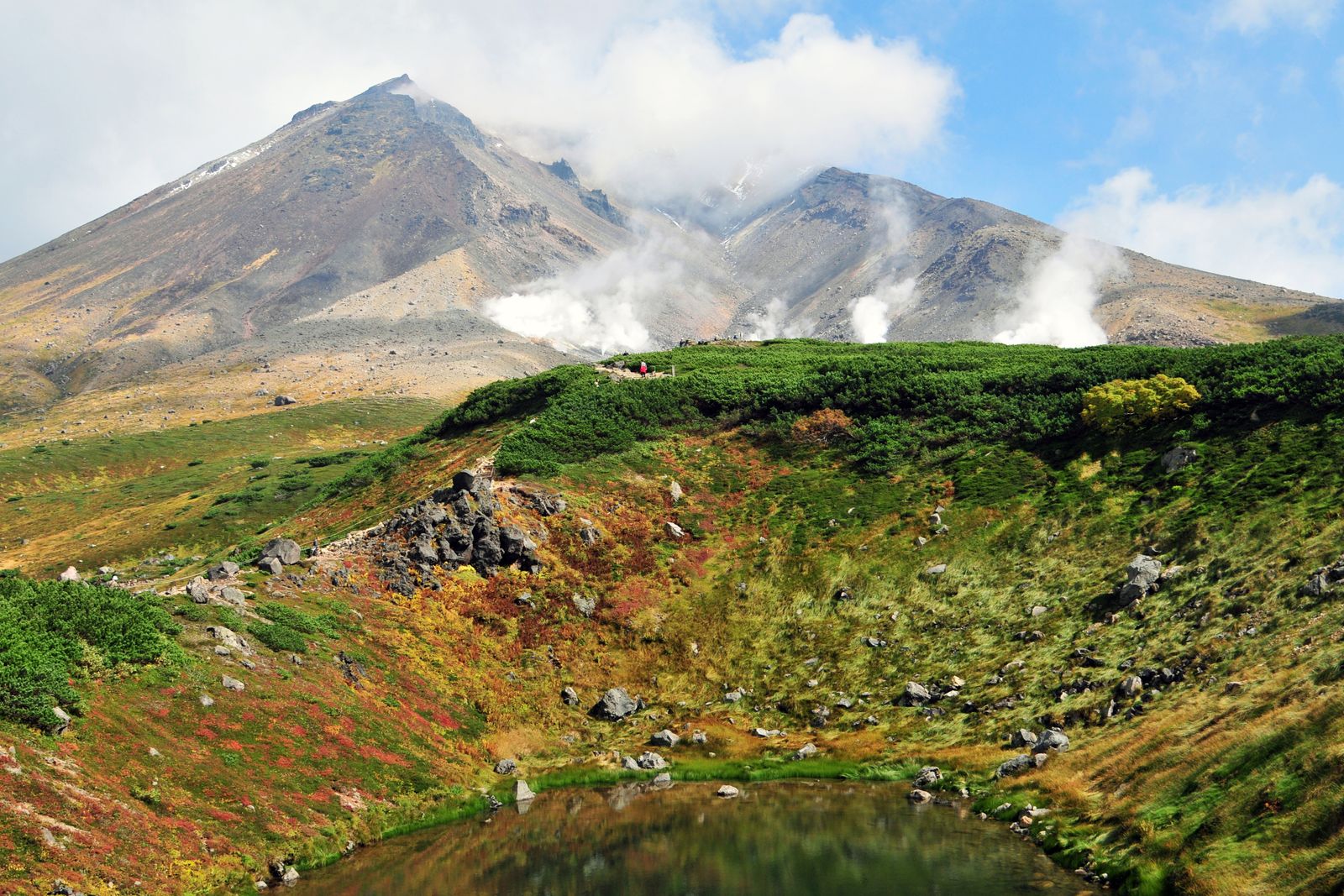 A steaming volcanic peak above a slope of red, orange and yellow autumn ground cover and a small still pond