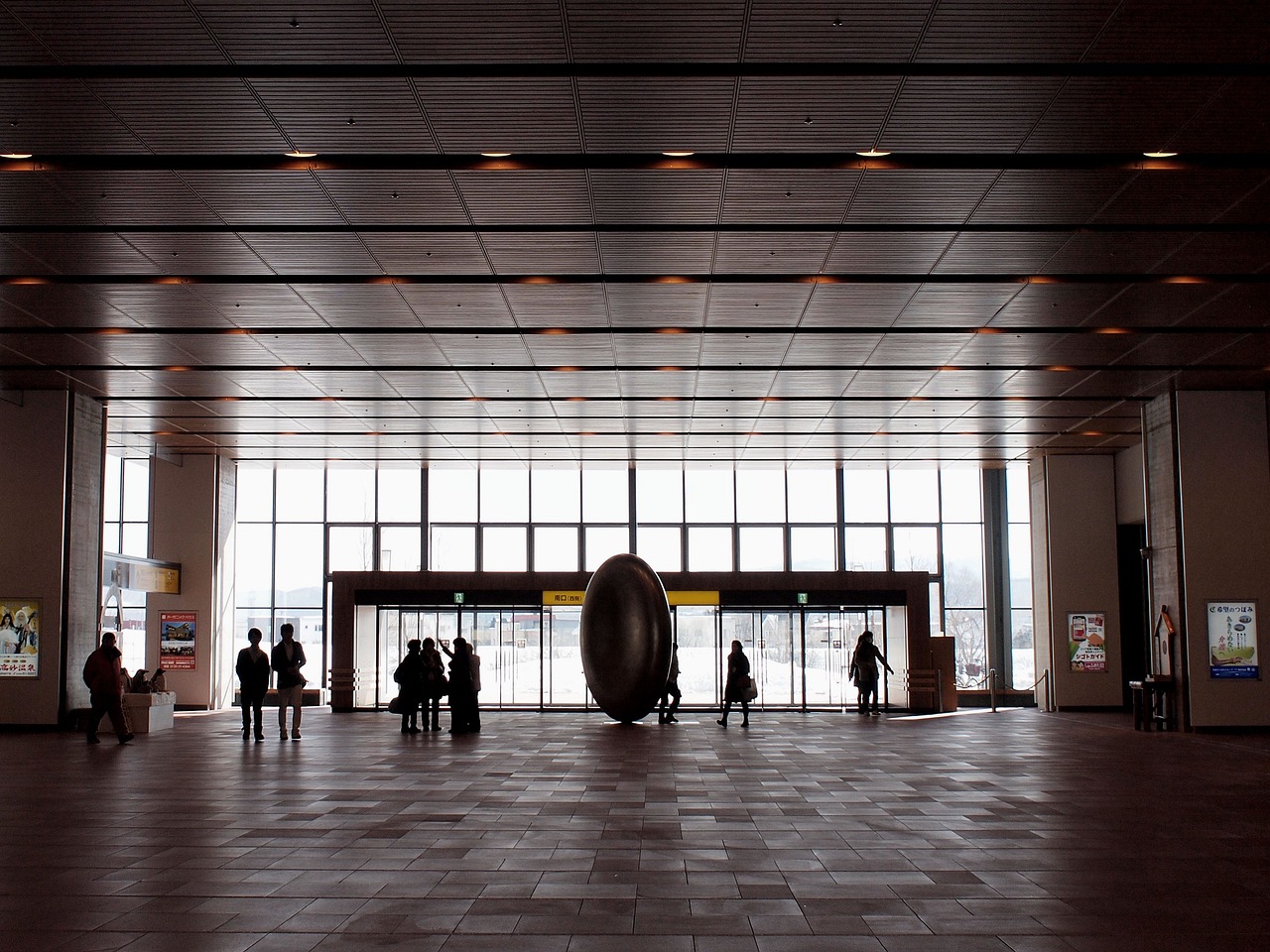 A station concourse with a large dark egg-shaped sculpture in front of the glass exit doors, snow on the ground outside