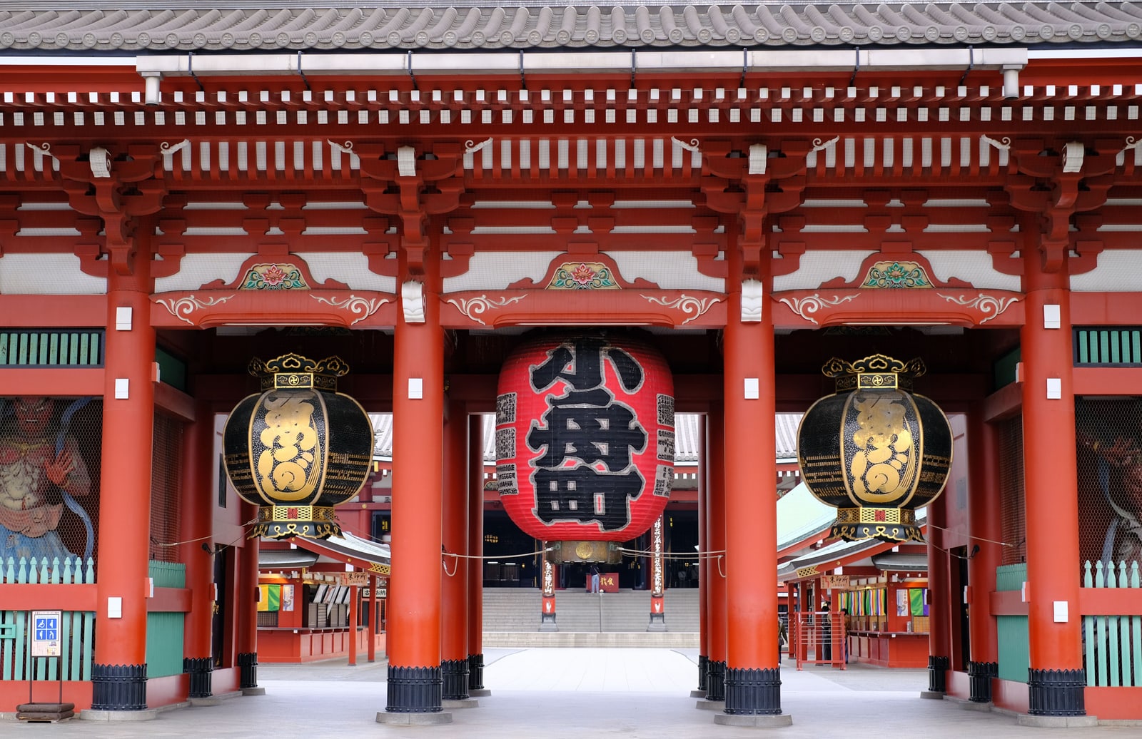 The Hozomon gate at Senso-ji, a large red lantern between two black-and-gold lanterns, guardian statues in the side niches and steps rising beyond