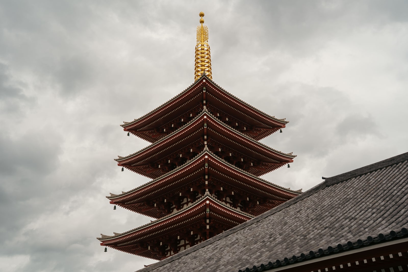 The upper tiers and gold finial of a five-storey red pagoda above a grey tile roof, under heavy cloud