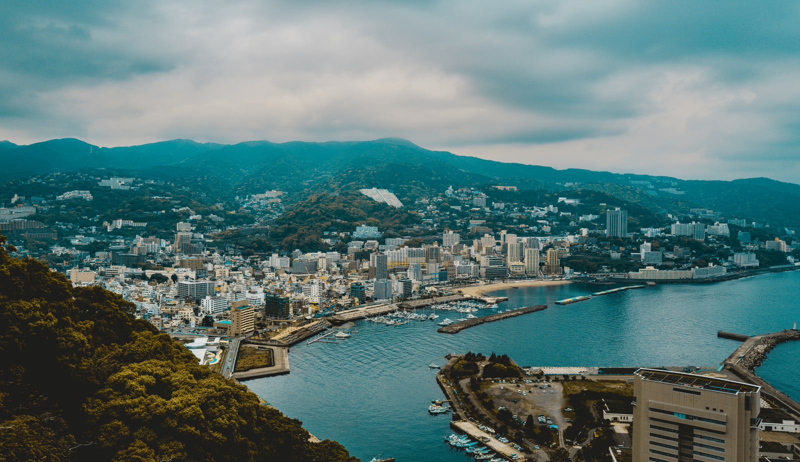 Atami town and bay from a hilltop under grey cloud: buildings on green hills, a curved sandy beach and a marina