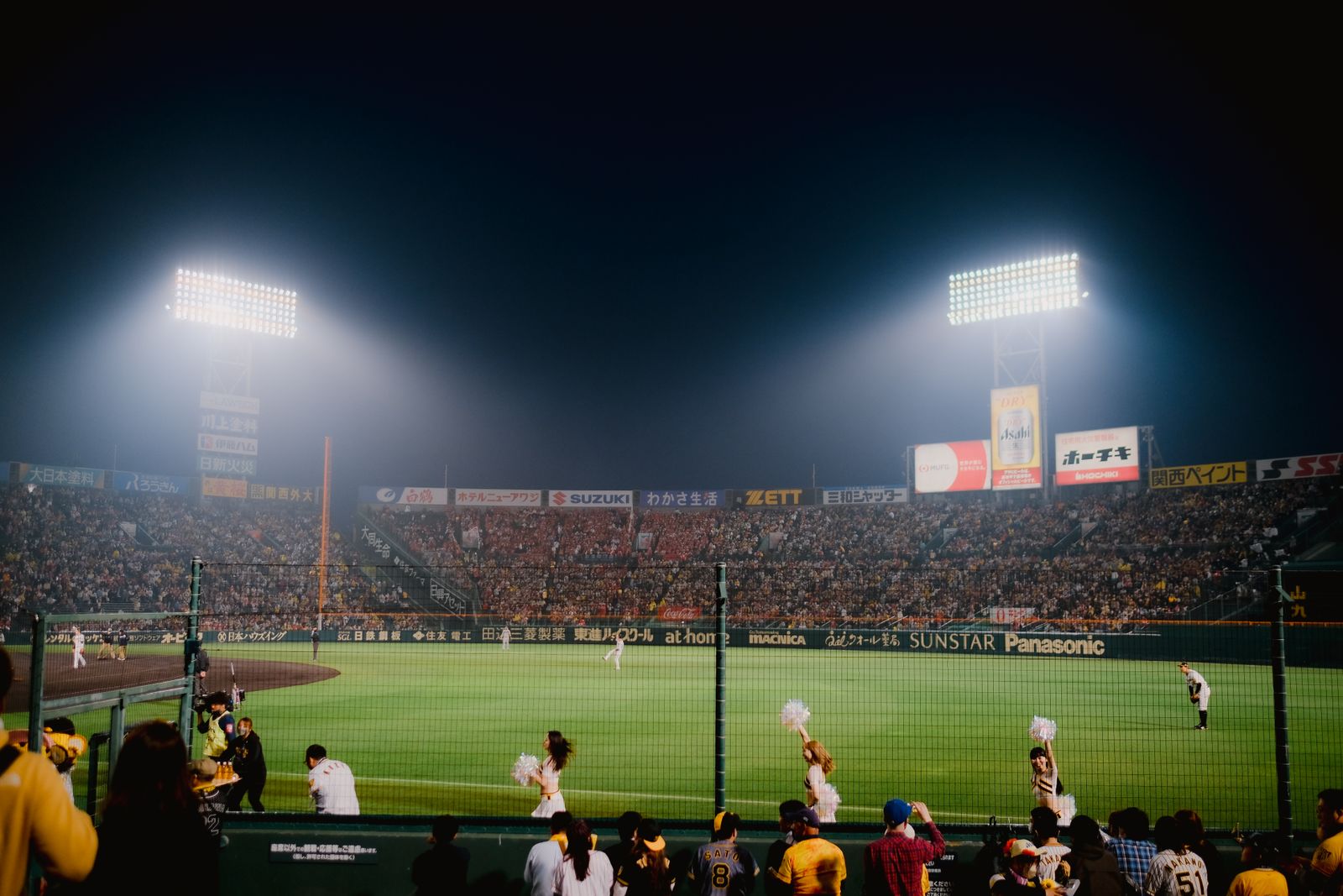 A packed open-air baseball stadium at night, floodlights on, cheerleaders on the outfield grass and fans in replica jerseys at the front rail.