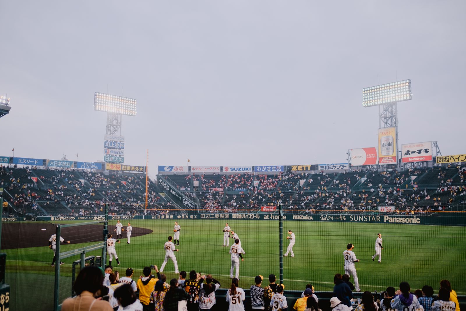 Uniformed players warming up in the outfield of a large stadium before a game, half-filled stands behind, fans in replica jerseys at the fence with phones out.