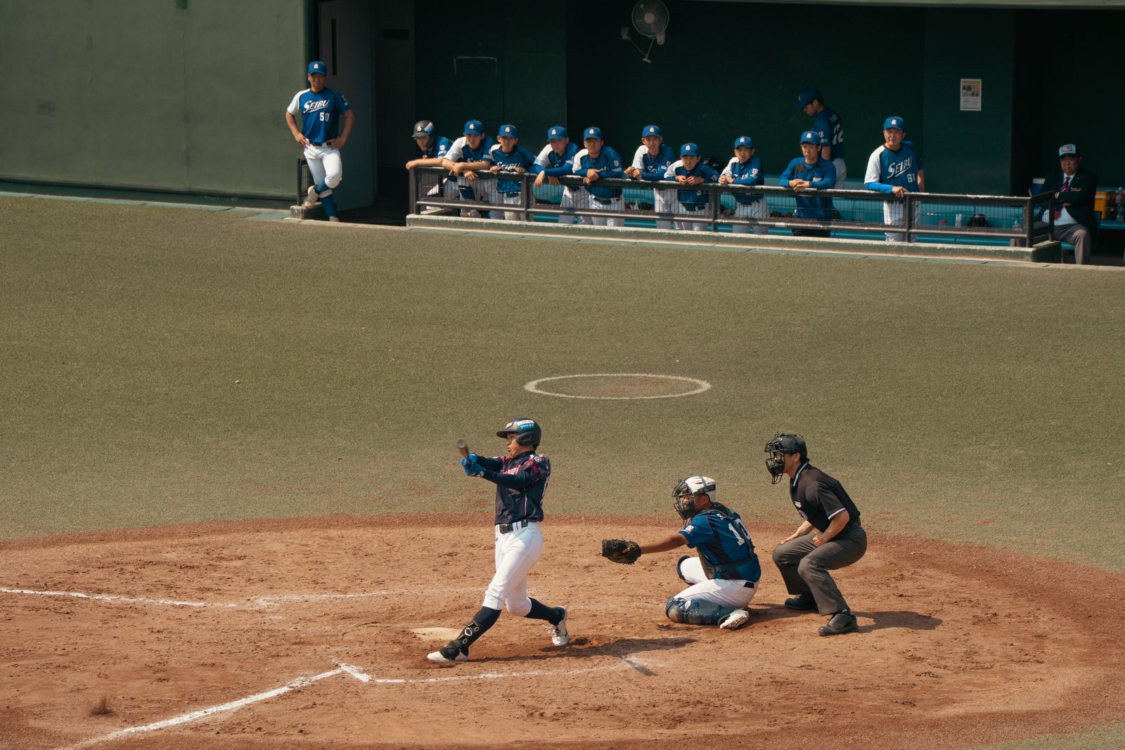 A batter finishing a swing at home plate with the catcher and umpire behind him, a dugout of teammates watching.