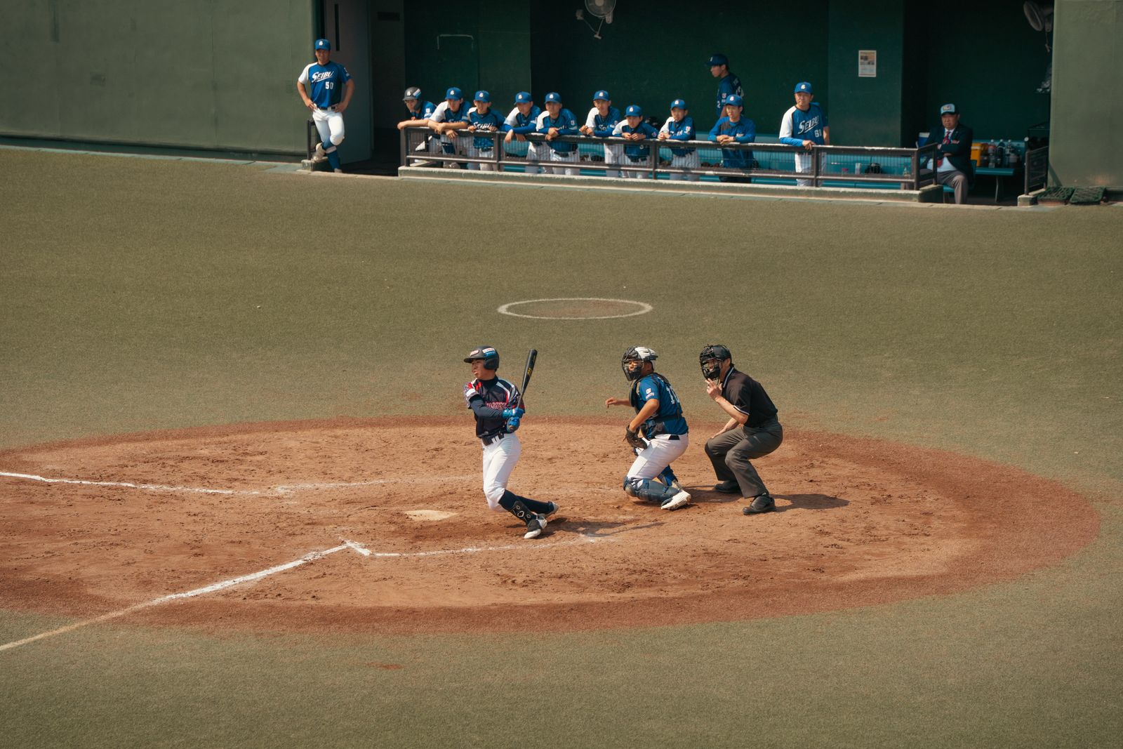 A batter loading his swing at home plate, catcher and umpire set, teammates at the dugout rail.