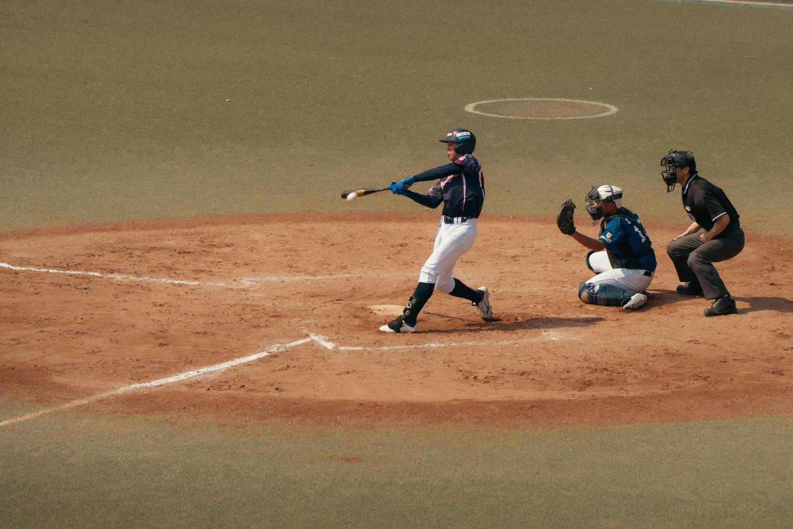 A batter making contact with the ball at home plate, catcher and umpire crouched behind.