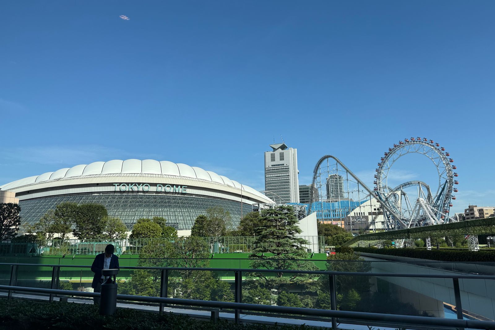 The white roof of Tokyo Dome under a clear blue sky, with a roller coaster and ferris wheel of the amusement park beside it.