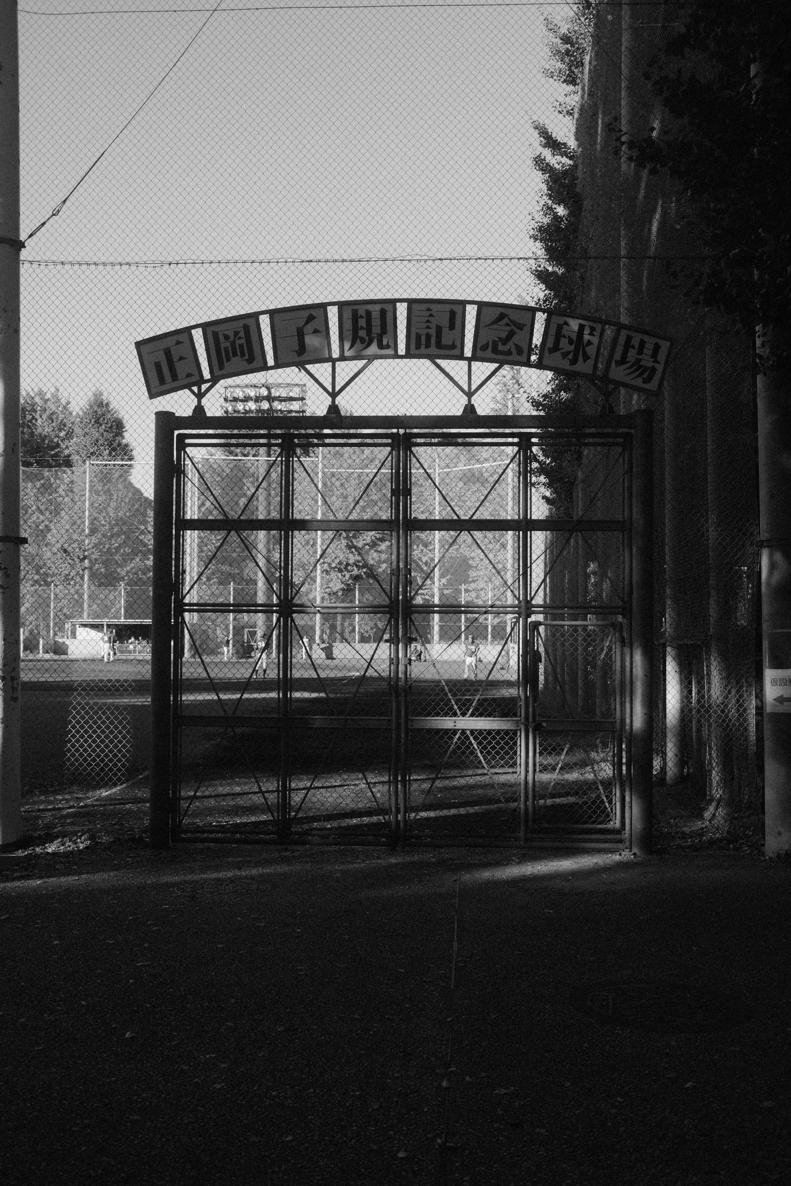 A closed metal gate to a public ballpark with a signboard above it, players visible on the field beyond, in black and white.