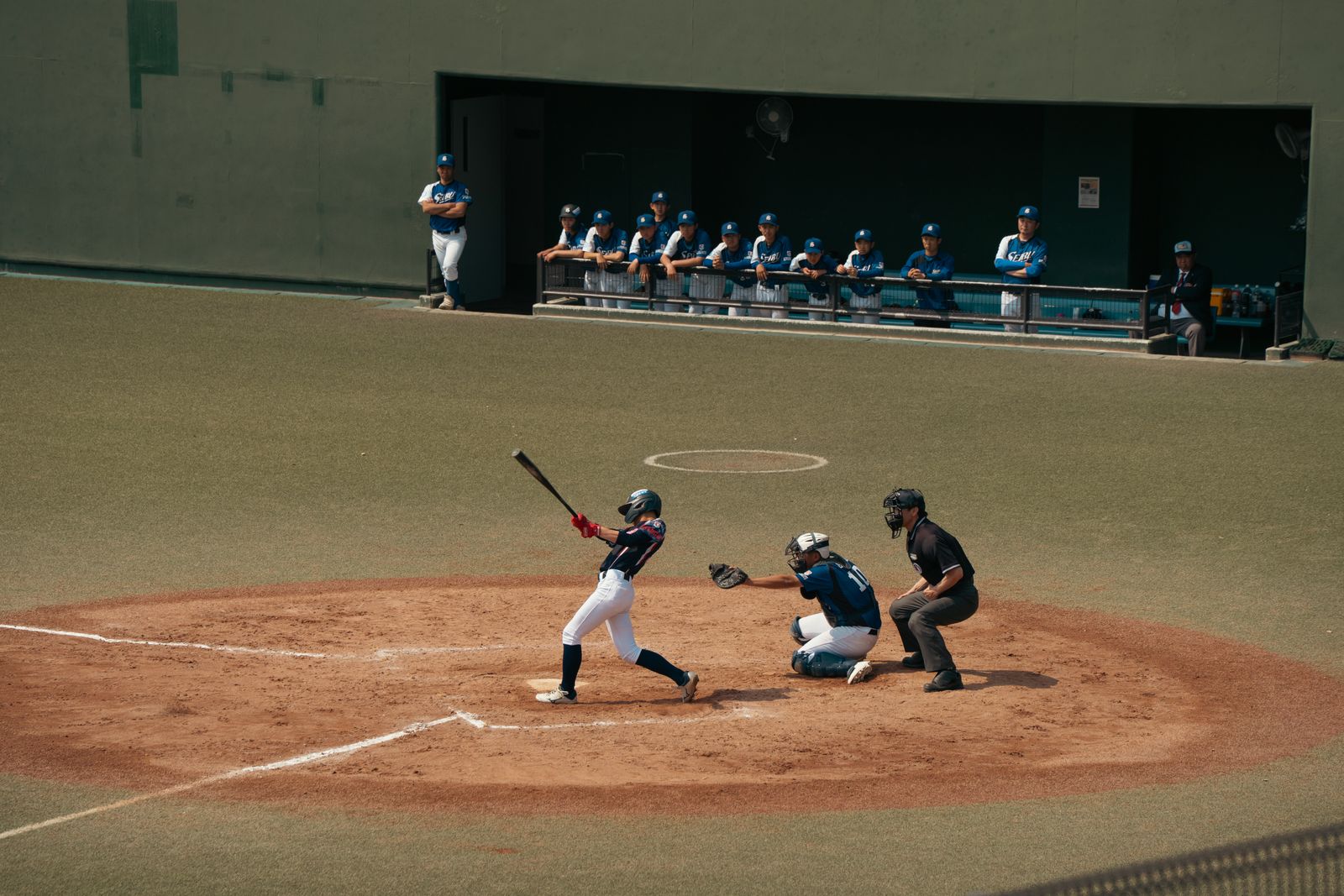 A batter beginning a swing at home plate, catcher and umpire behind, a dugout of teammates leaning on the rail.