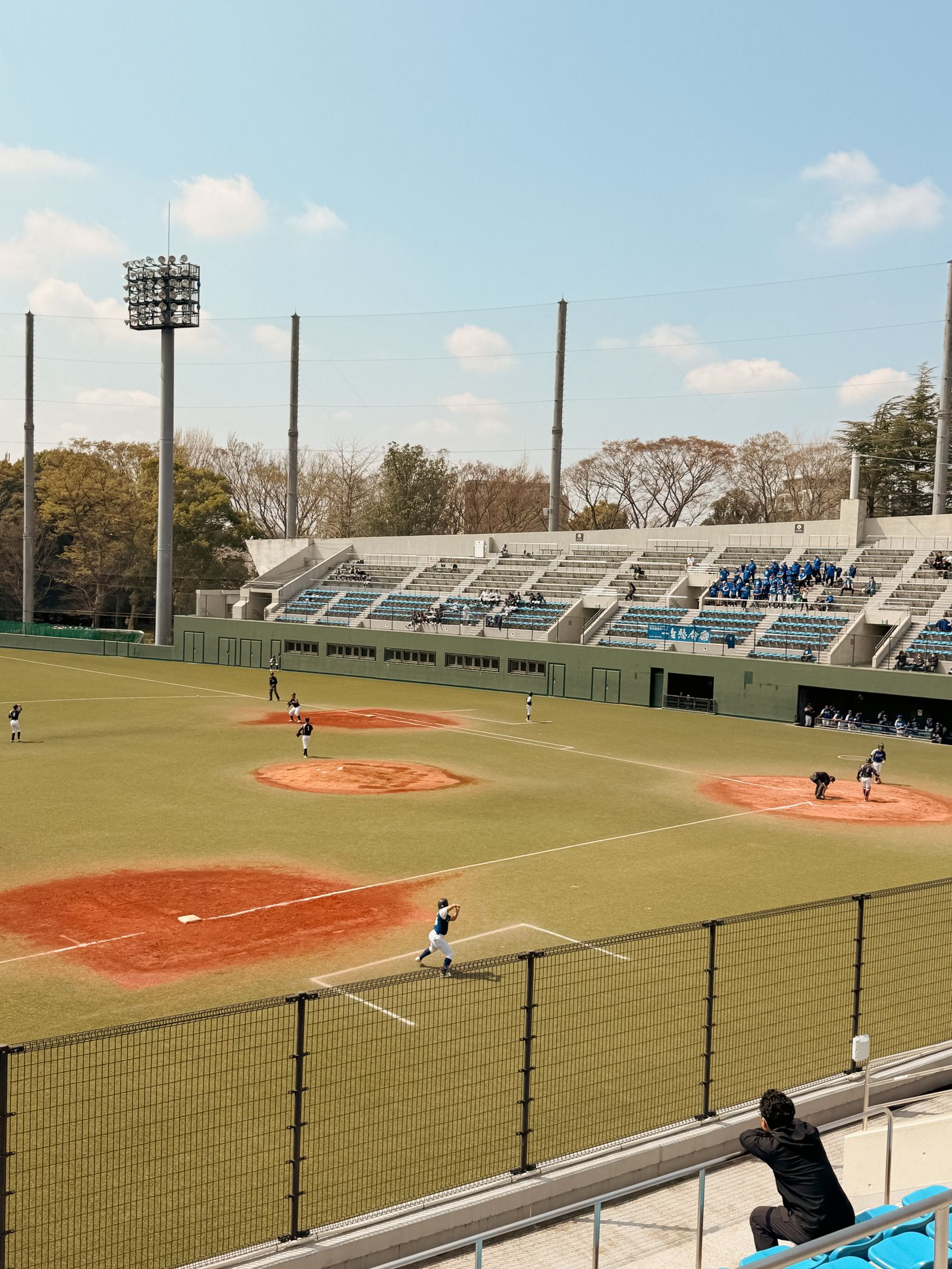 A daytime amateur baseball game on an artificial-turf field at a small ballpark, a small stand of blue seats behind, seen over a fence from the stands.
