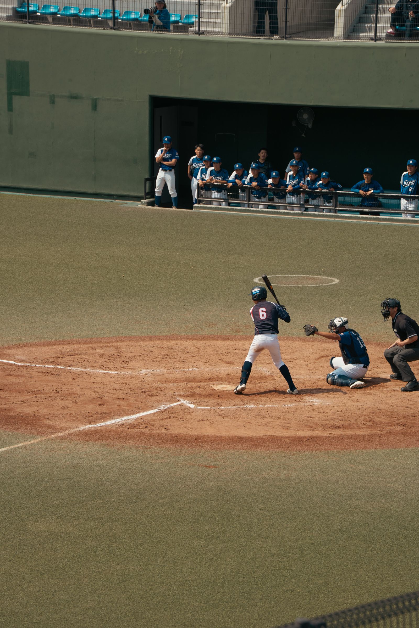 A batter, catcher and umpire at home plate on a dirt cutout, teammates watching from a dugout behind, at a small ballpark.
