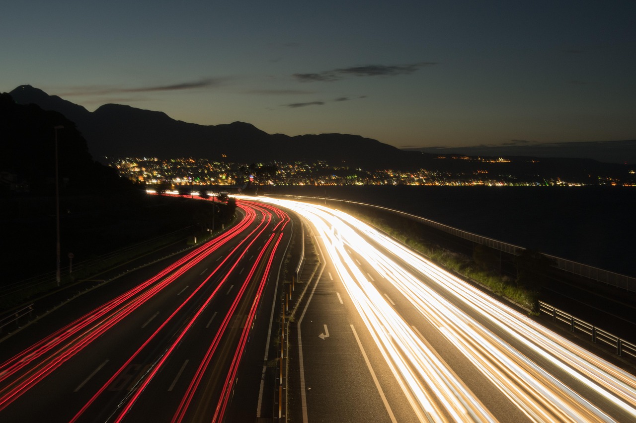 Light trails on a coastal road at dusk, with town lights along a bay below the mountains