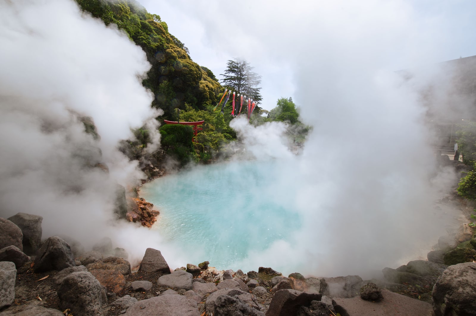 A steaming cobalt-blue hot spring pond ringed by rocks, a small red torii and carp streamers on the slope behind