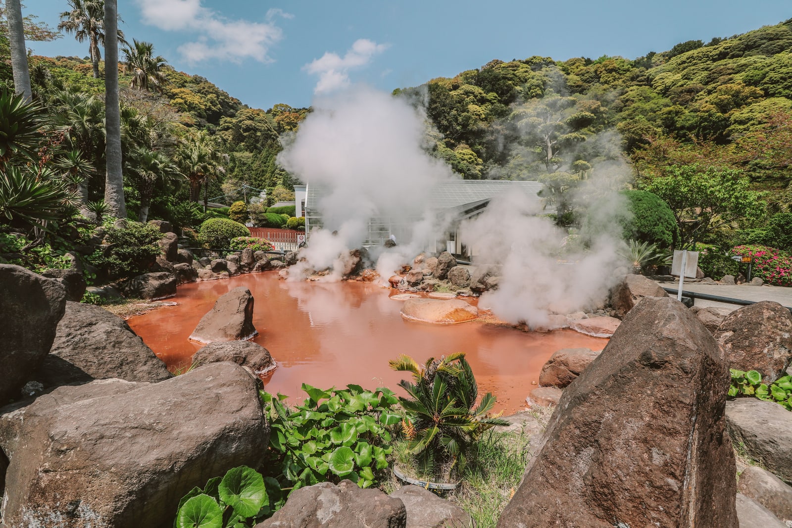 A steaming red-orange hot spring pond among large rocks and palms under a blue sky