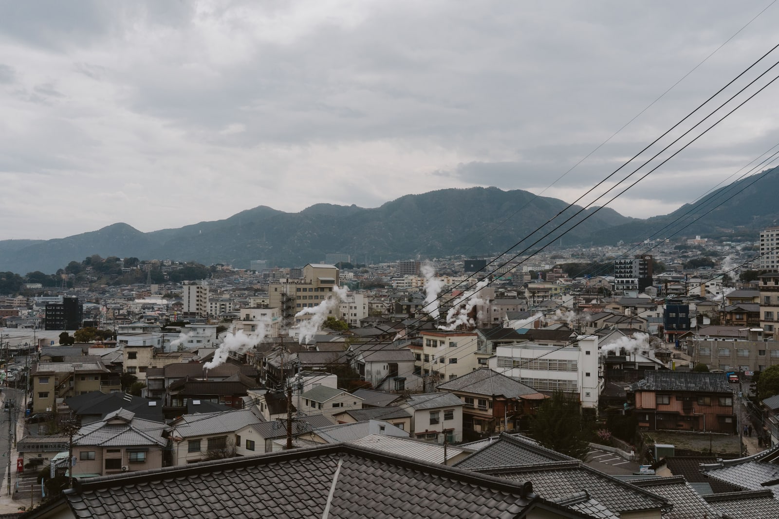 White steam rising from vents between the tiled rooftops of a hillside town, a forested ridge behind