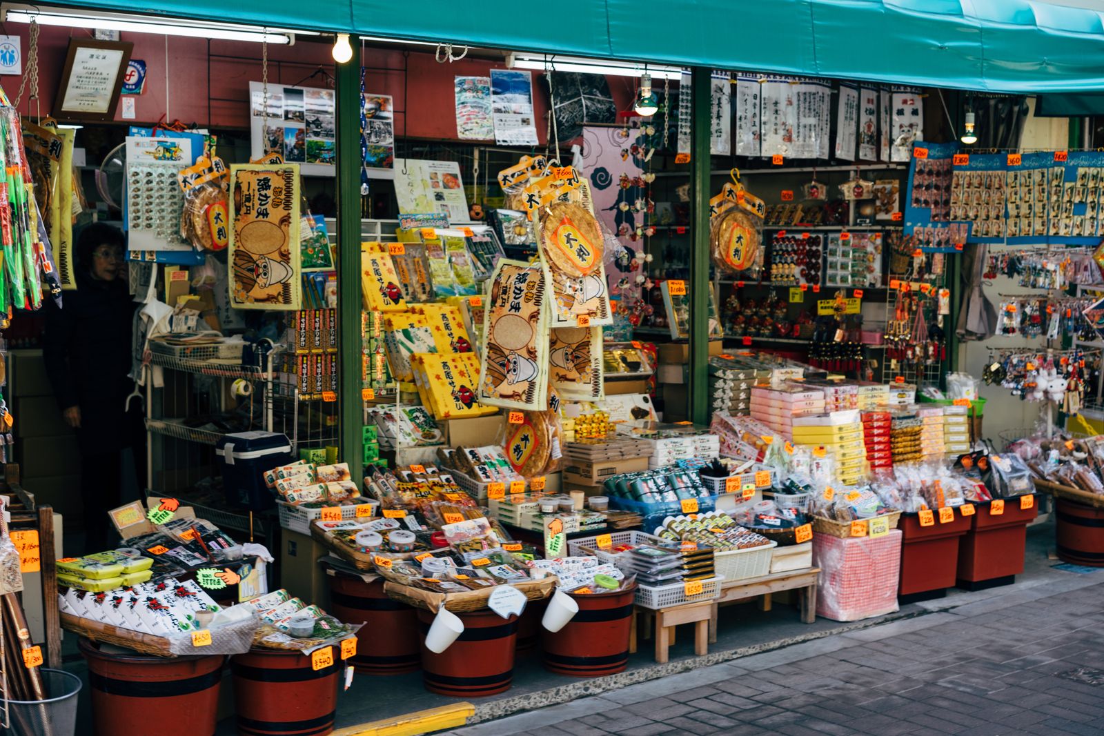 An open souvenir shopfront under a teal awning, boxed confectionery and oversized senbei on strings, yen price cards on everything.