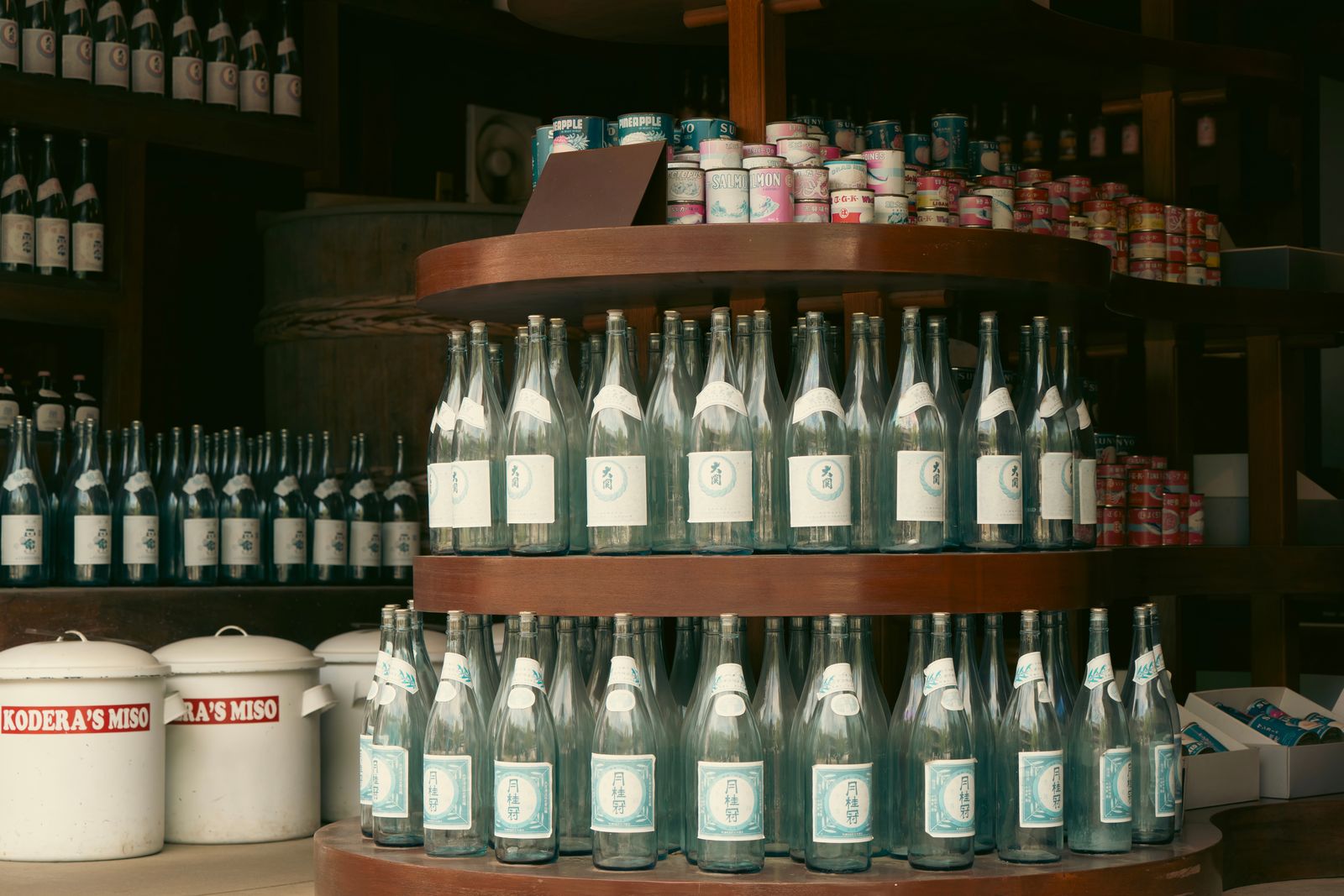 Tiers of large sake bottles with 大関 and 月桂冠 labels in a wooden shop display, vintage canned goods on the shelf above.