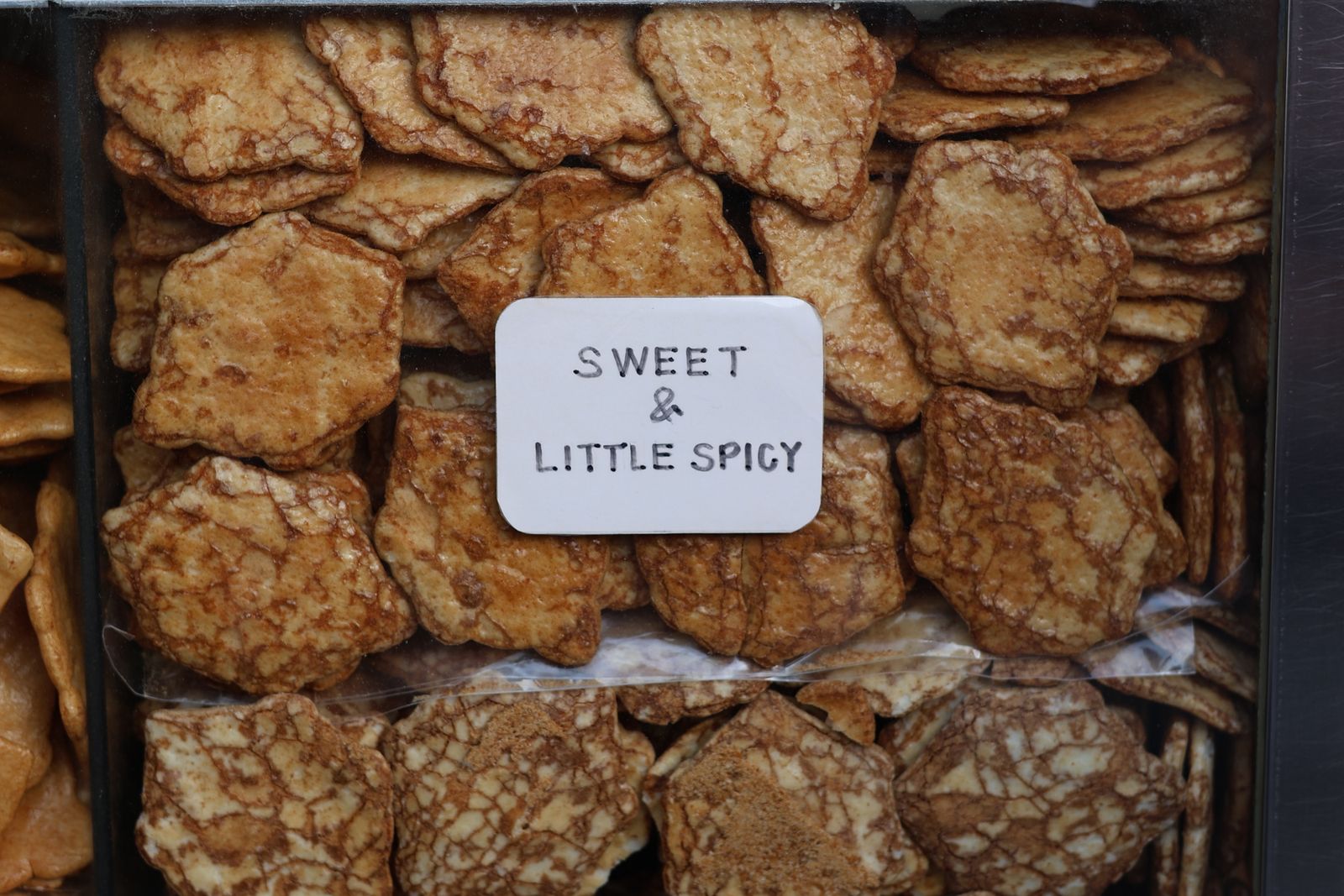 Glazed senbei rice crackers heaped in a glass-fronted shop case, a row of bagged ones beneath, and a handwritten card reading SWEET & LITTLE SPICY.