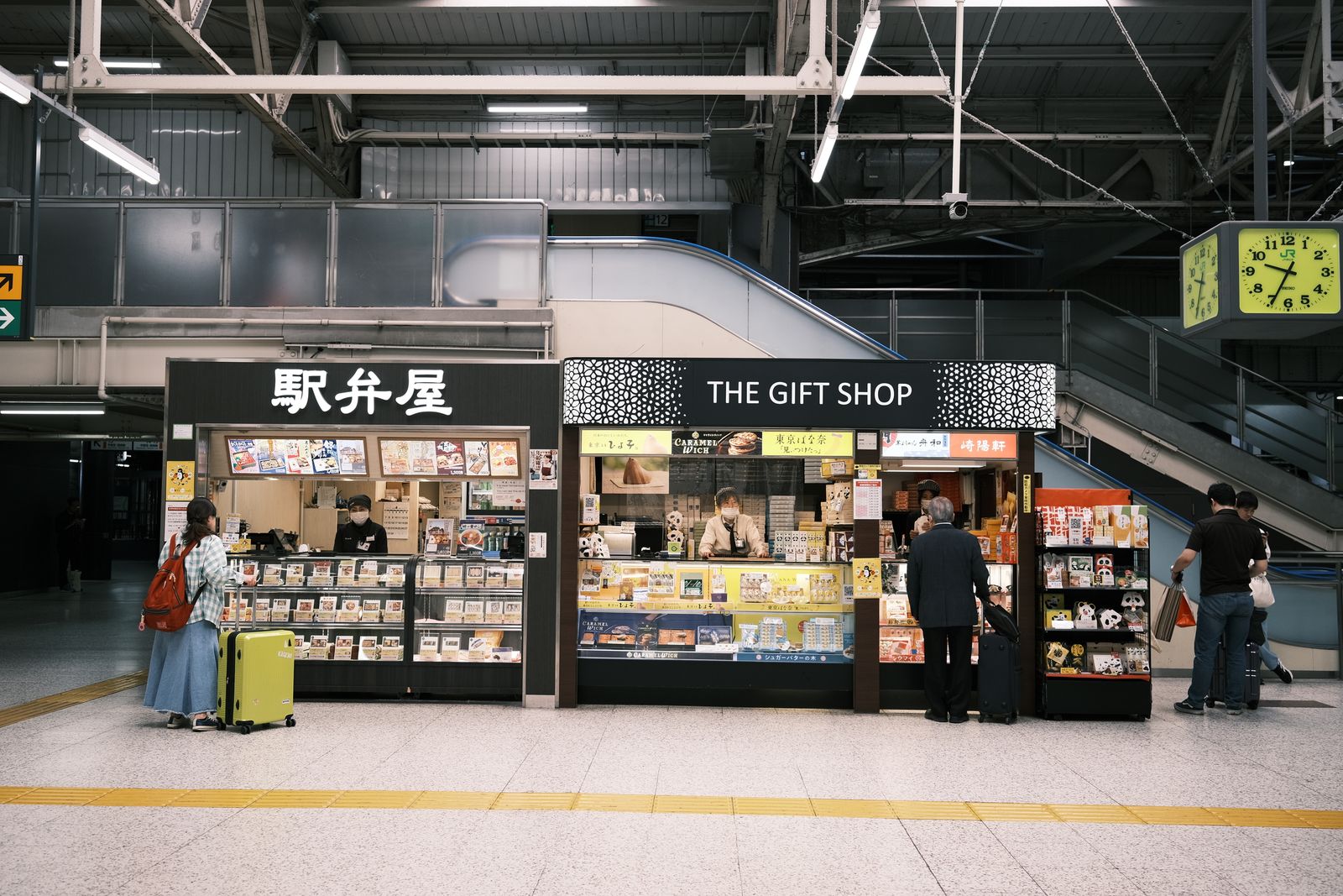A JR station concourse with an ekiben stall signed 駅弁屋 beside a kiosk signed THE GIFT SHOP, its header carrying Tokyo Banana, ひよ子, Caramel Wich and 崎陽軒; two travelers face the counters with their backs to the camera.