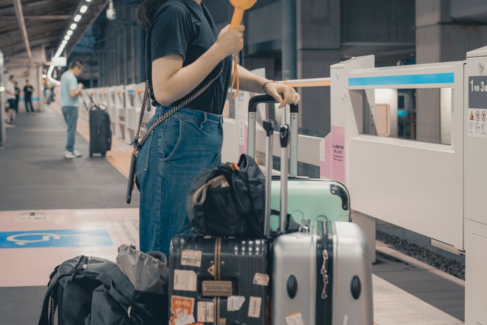 A traveler in denim, seen from the shoulders down, beside a stack of three suitcases and two backpacks on a Japanese station platform with half-height platform doors.