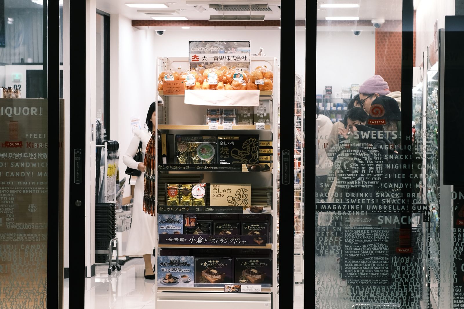 The glass entrance of a station-building convenience store at night, a rack of boxed Nagoya-region sweets — 金しゃち焼チョコ and 小倉トーストラングドシャ — just inside the doors, and bagged mikan on the top shelf.
