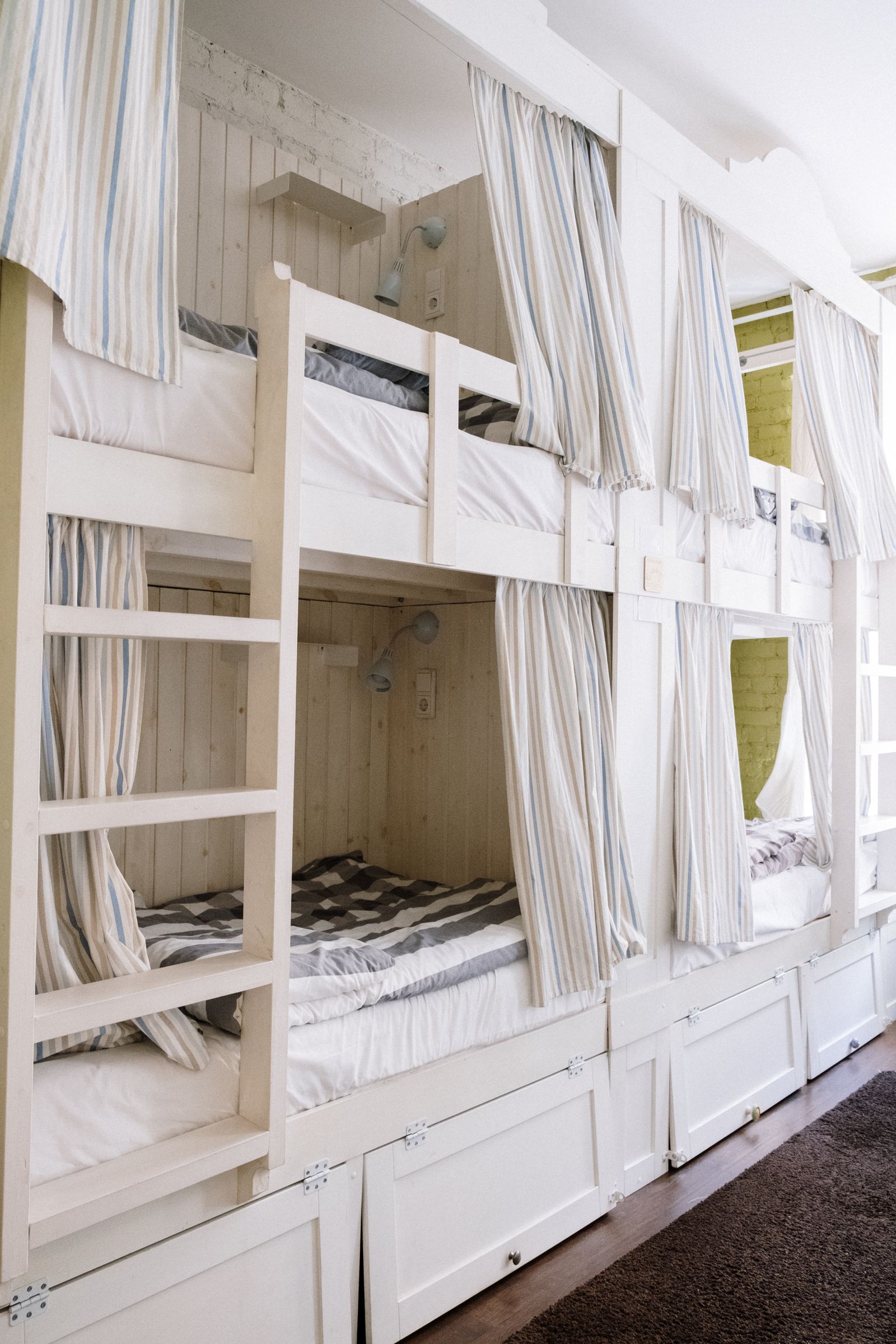 Hostel dorm bunks built into white timber boxes, each with a striped privacy curtain, a reading lamp and a drawer beneath.