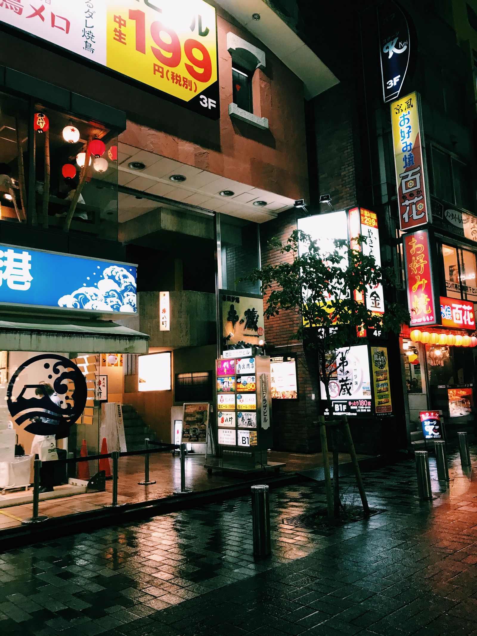 A rain-wet pavement at night lined with lit izakaya signs and red lanterns, almost empty.