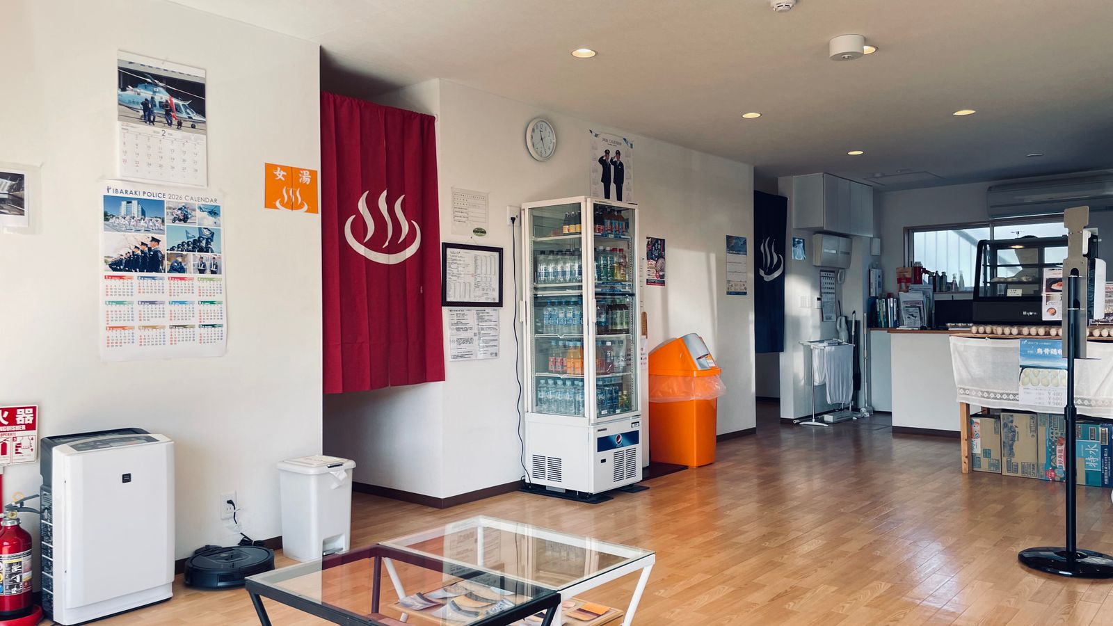 The reception lounge of a small Japanese bathhouse: a red noren with the hot-spring mark hangs in the doorway to the women's bath, a drinks fridge beside it.