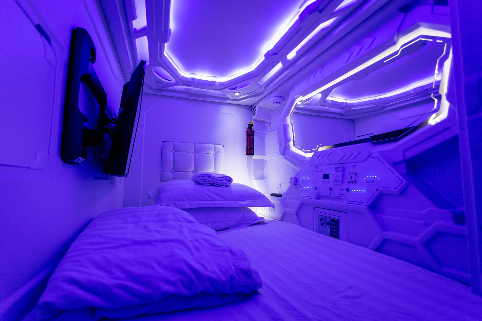 Inside a capsule hotel pod: a single bed, a wall-mounted screen and a control panel under blue LED lighting.