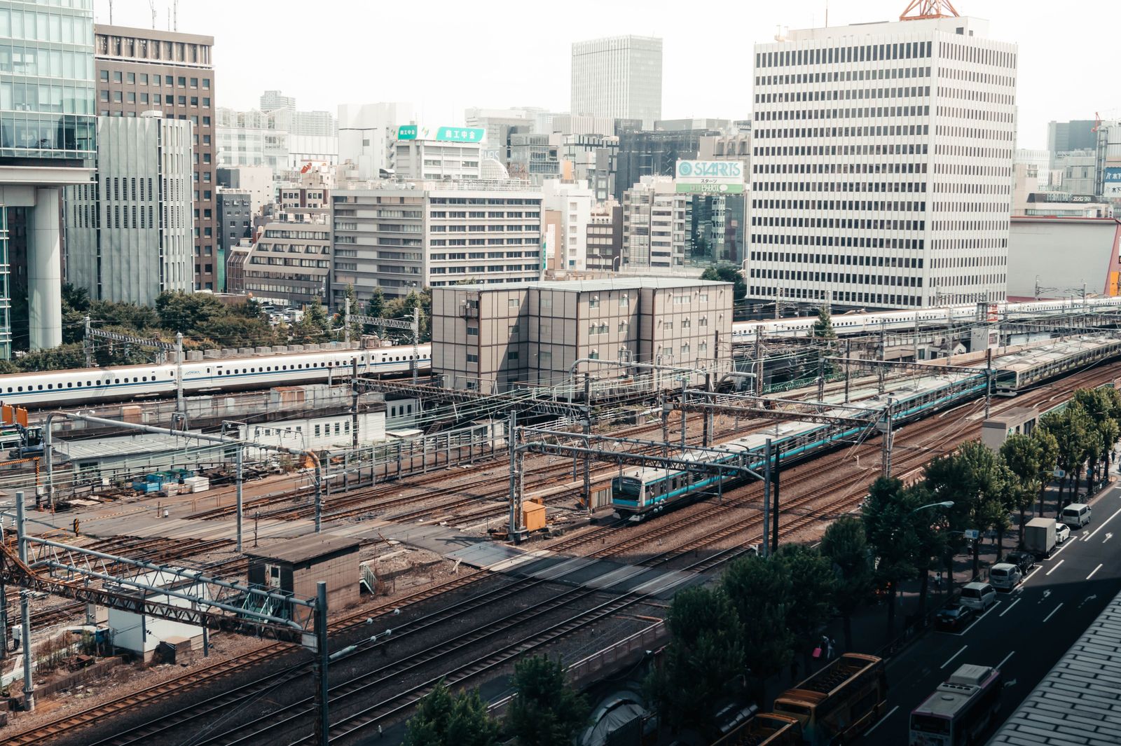 Daytime view over the tracks beside Tokyo Station: a Shinkansen on the far line, a local train in the middle, office towers behind.