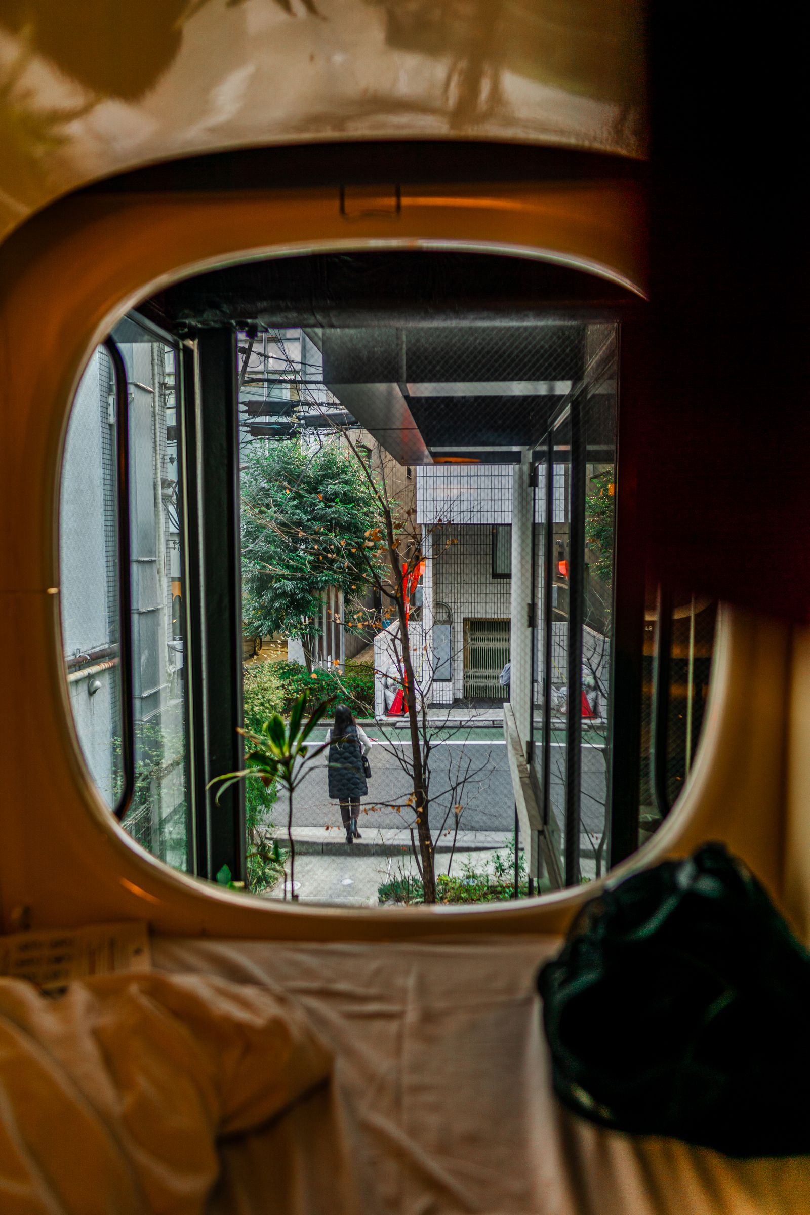 Looking out of a capsule pod's rounded window onto a quiet city street in the morning; one person walks away down the pavement.