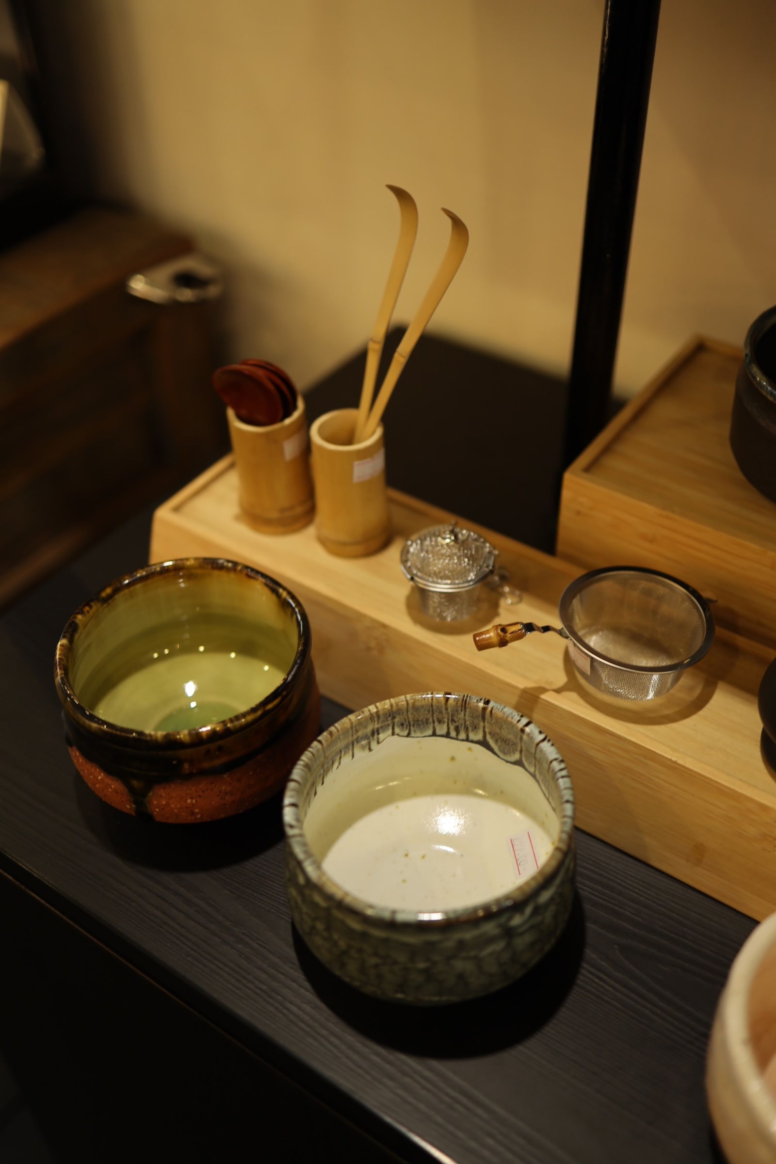 Ceramic tea bowls, bamboo tea utensils, and a metal tea strainer arranged on a wooden stand.