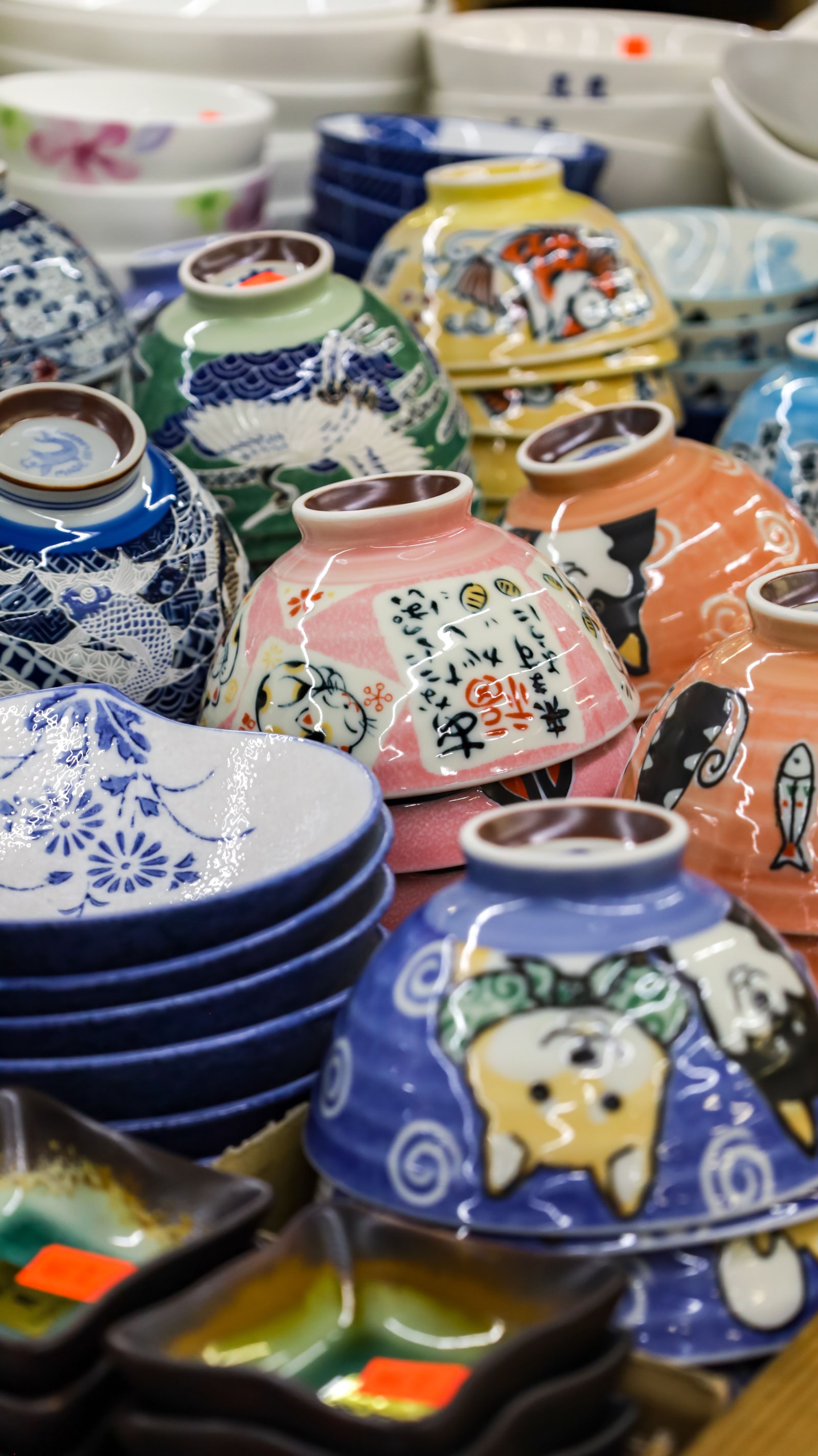 A dense stack of colorful Japanese-style ceramic bowls and small vases with painted motifs (cranes, cats, fish, floral) on a shop shelf/table.