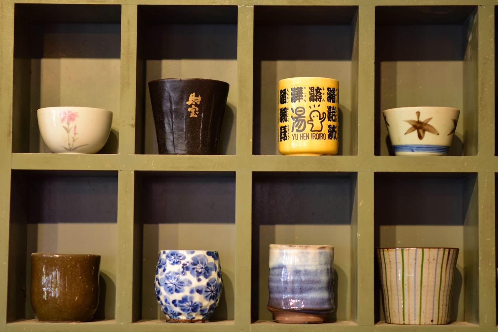 Eight assorted ceramic cups of different glazes and patterns displayed in a wooden cubby-hole shelf.