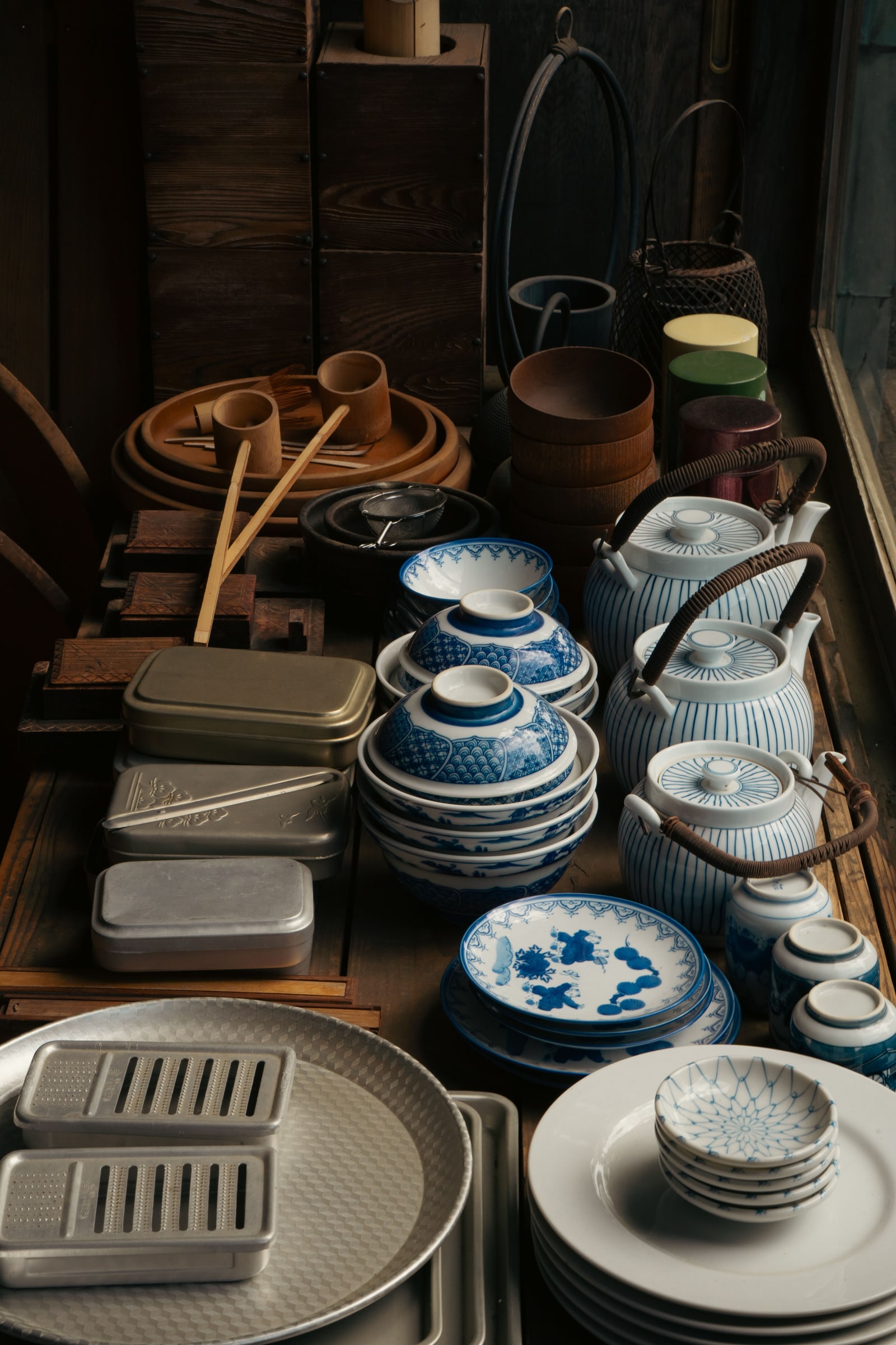 Traditional Japanese pottery and dishware — bowls, jugs, a woven tray, vases — arranged on a dark wooden shelf near a window.