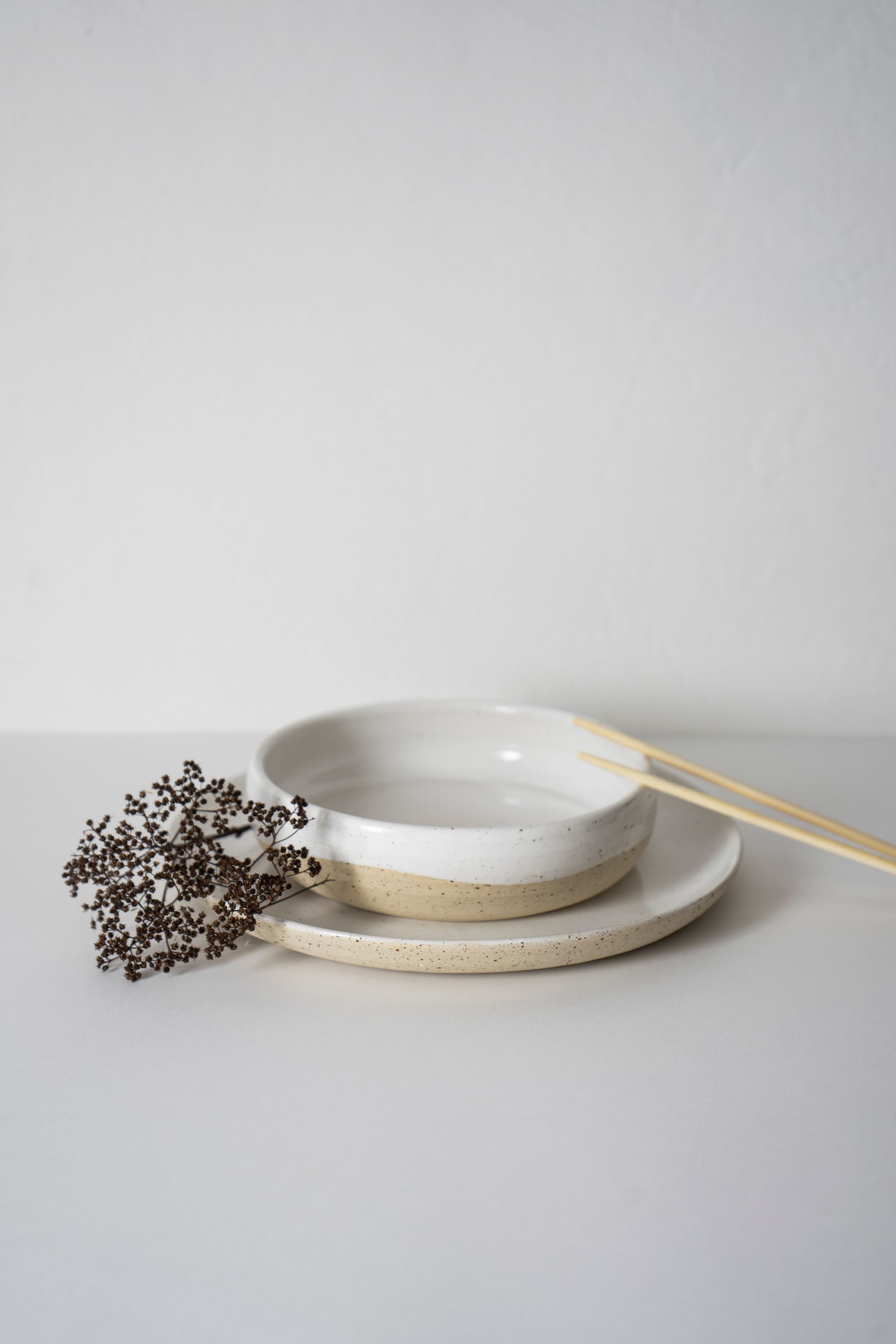 A plain white ceramic bowl with a pair of chopsticks resting on it, on a plain light background.