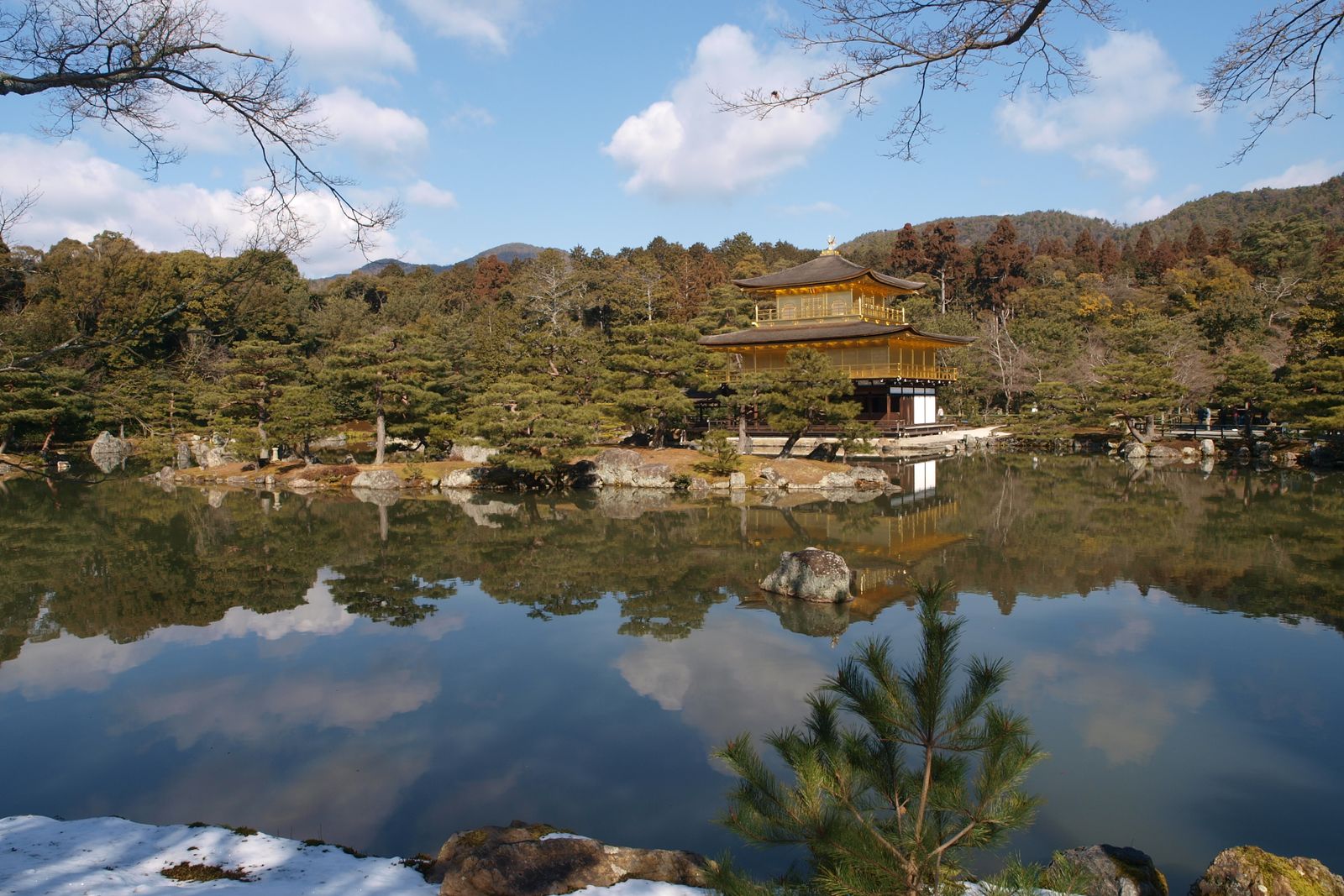 Kinkaku-ji reflected in its pond under winter light, snow lingering on the foreground rocks.