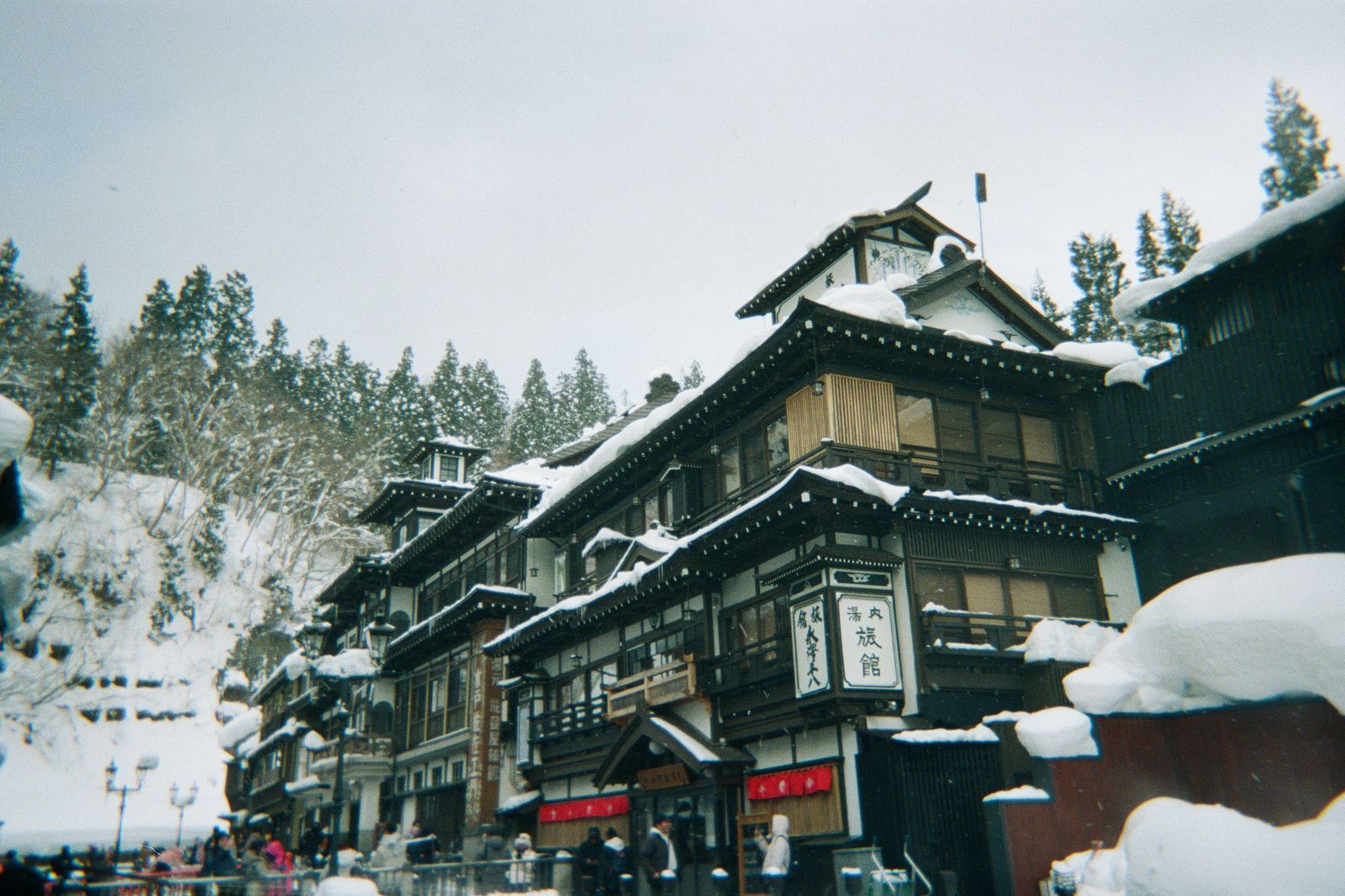 A snow-country onsen street lined with old ryokan under heavy snow, visitors small in the lane below.